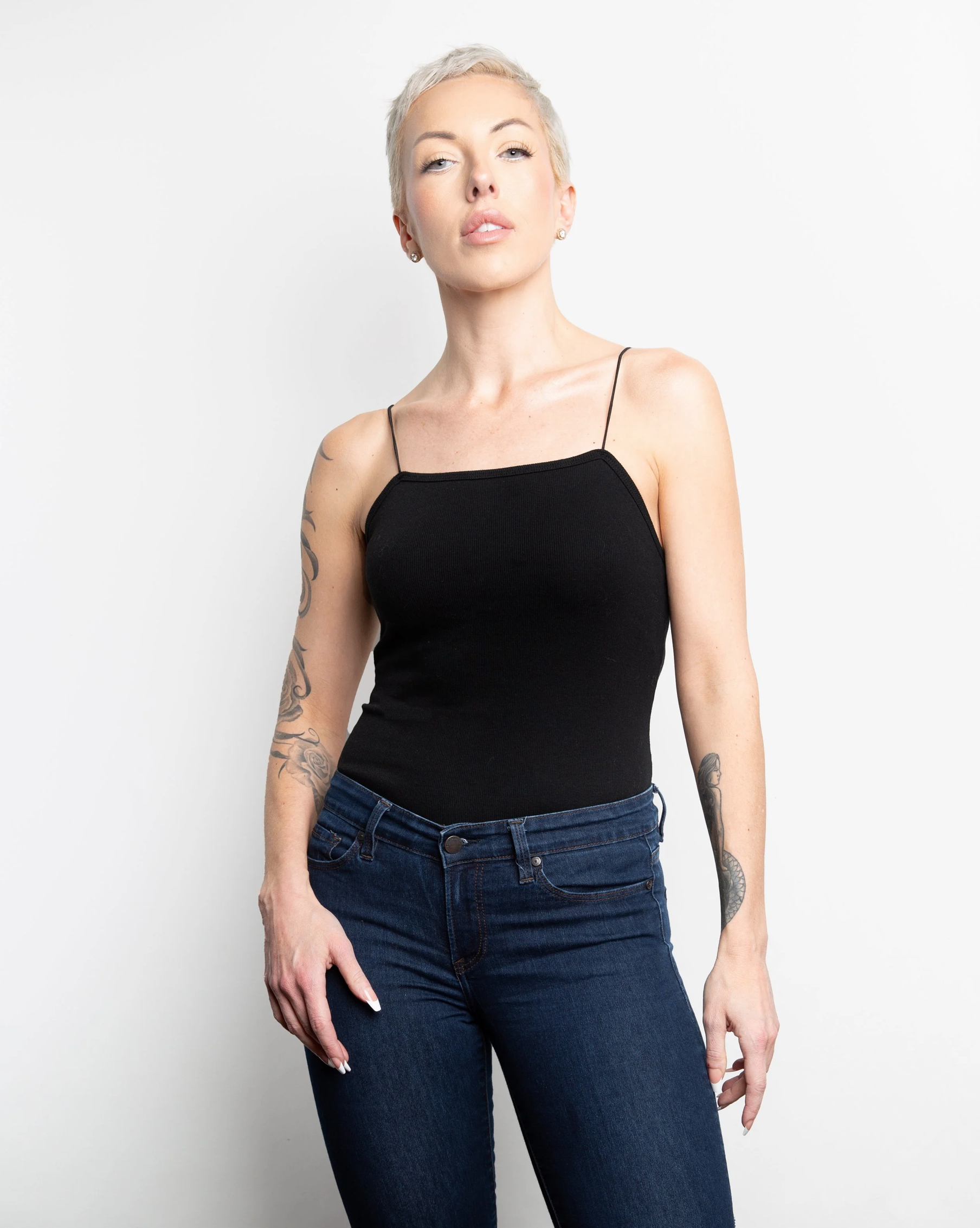 Studio portrait of a model with short blonde hair and tattooed arms wearing a black sleeveless top and dark blue jeans. She stands confidently against a clean white background, looking directly at the camera.