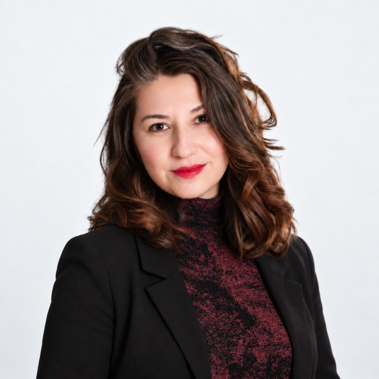 Headshot of a young woman with curly brown hair, wearing a black blazer and a red patterned turtleneck, smiling softly against a plain white background.