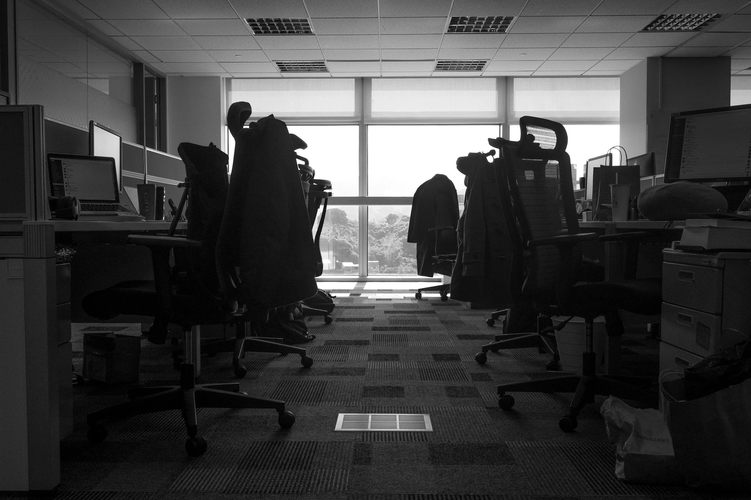 Empty office workspace with desks, office chairs, monitors, and bags, facing large windows with a view of trees outside.