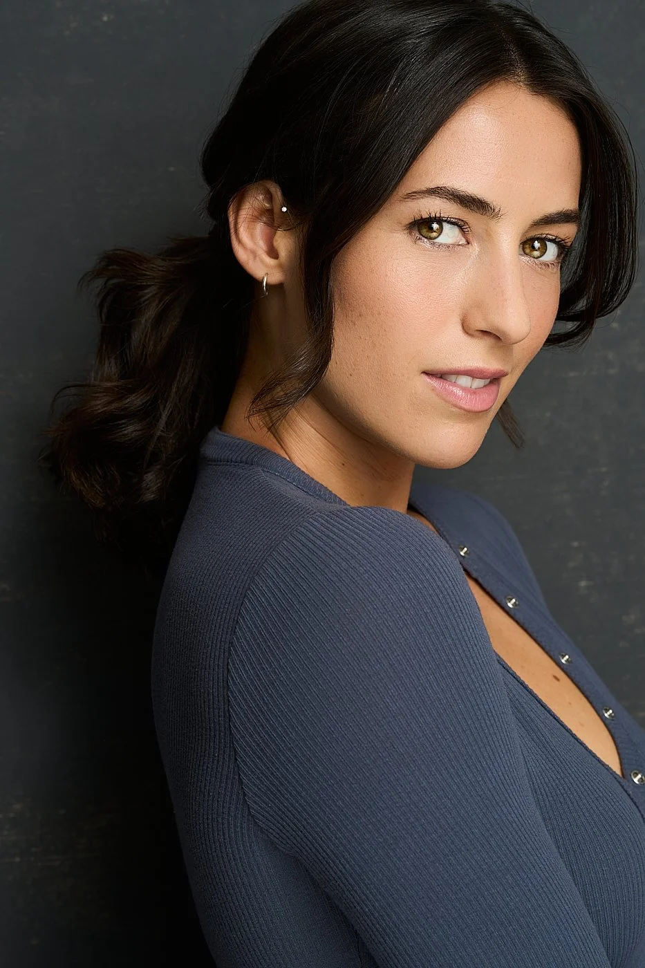 Close-up of a woman with dark wavy hair, wearing earrings, and a blue top with metallic buttons, looking at the camera against a dark background.