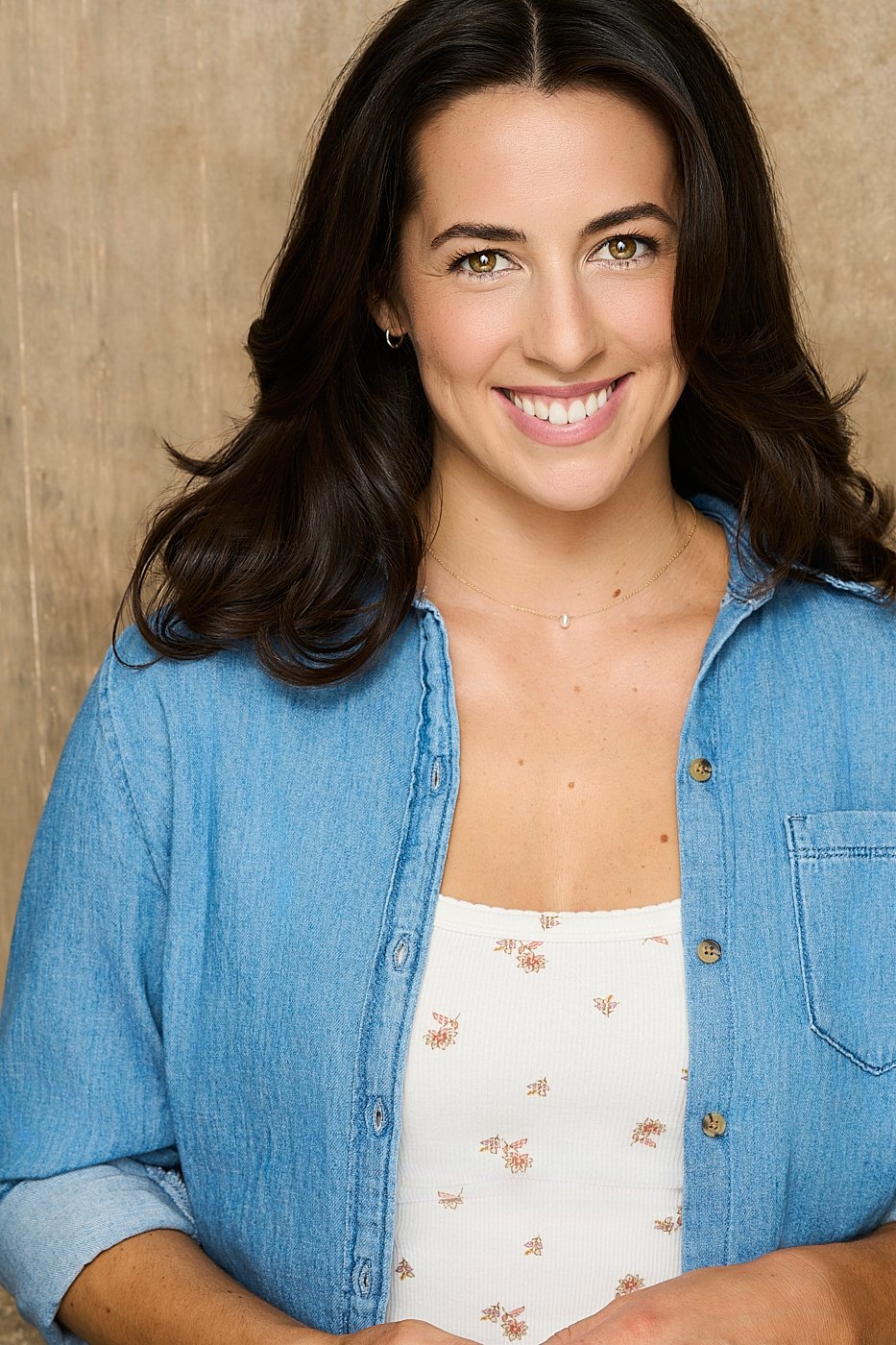A smiling woman with long dark hair wearing a denim shirt over a white floral top, standing in front of a wooden background.