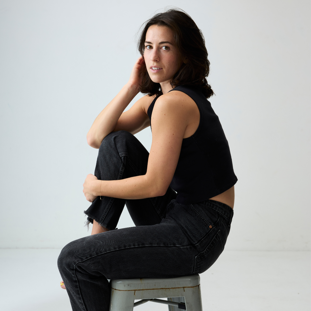 A young woman with dark hair, wearing a black sleeveless top and black jeans, sitting on a small metal stool against a plain white background.