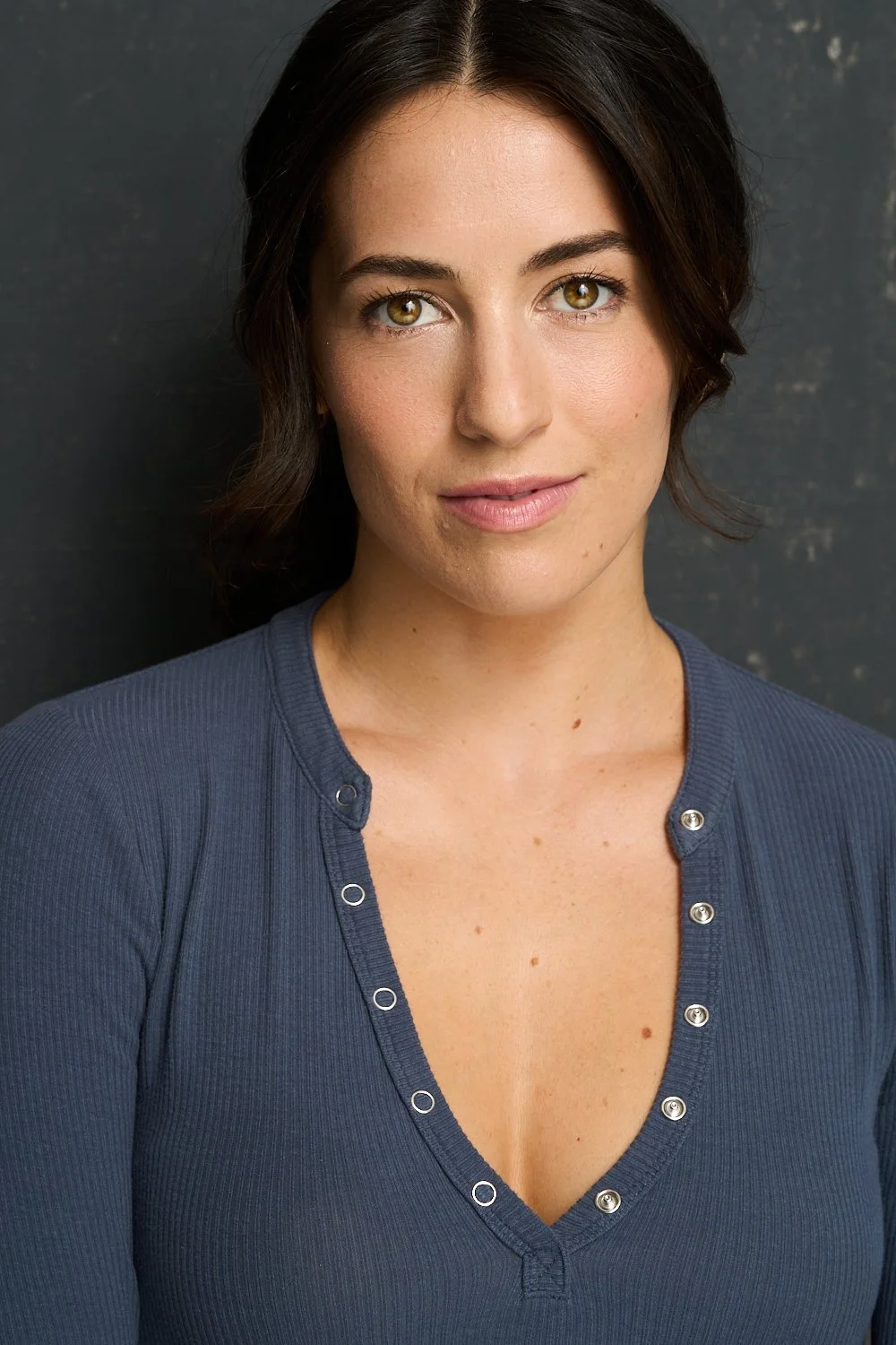 Close-up of a woman with dark brown hair, hazel eyes, and light skin, wearing a navy blue top with metallic buttons, standing against a dark textured background.