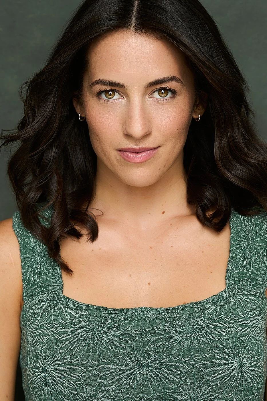 Close-up portrait of a woman with wavy dark hair, wearing a green textured sleeveless top, smiling softly, against a plain background.