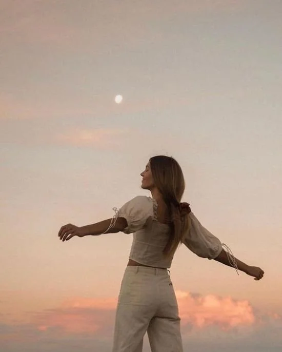 A woman standing outdoors at sunset with her arms outstretched, facing sideways, under a sky with a visible moon and pink clouds.
