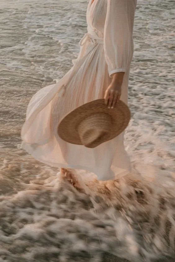 Person wearing a light-colored flowing dress walking in the water at the beach, holding a straw hat.