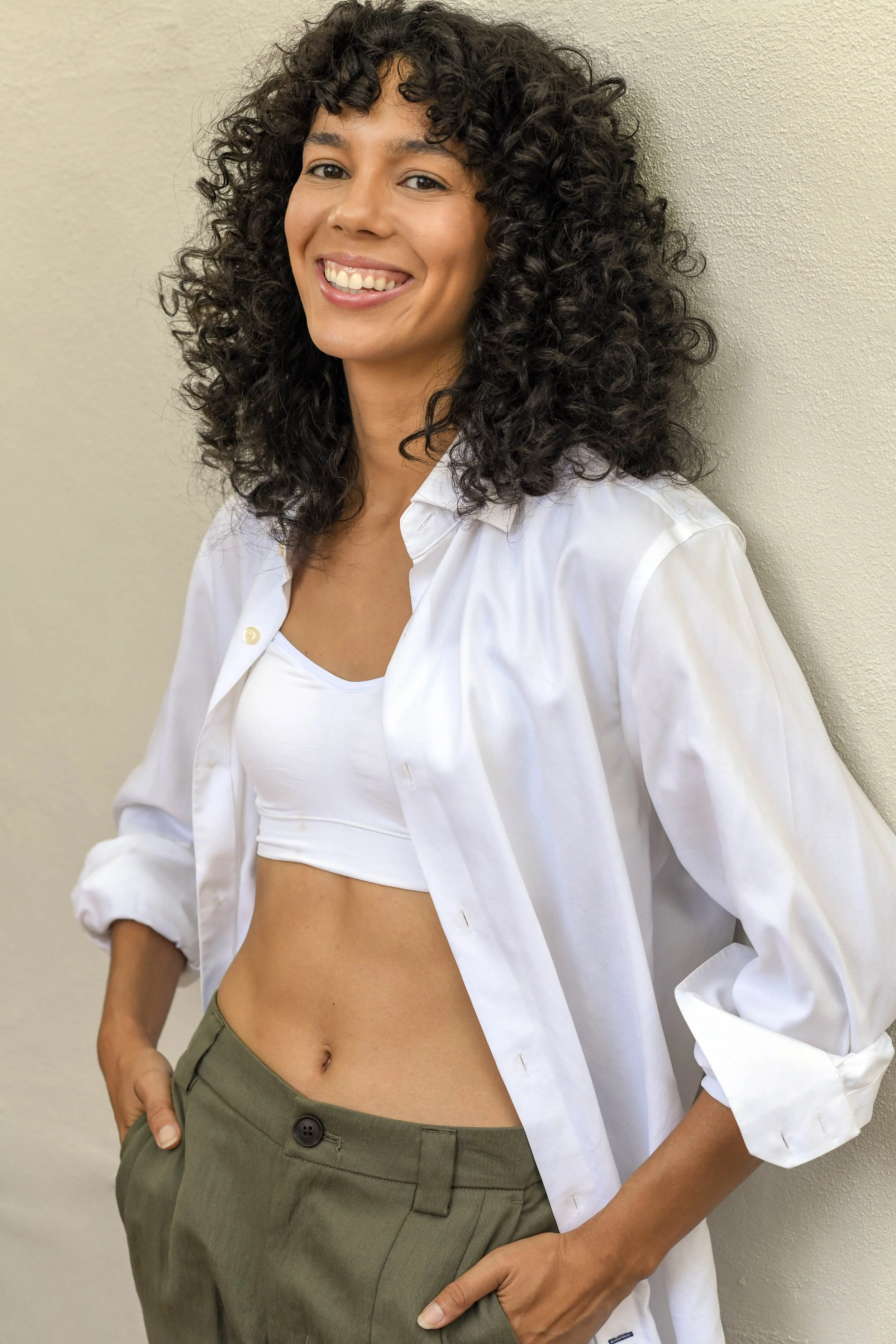 A smiling woman with curly dark hair, wearing a white open shirt over a white crop top and olive green pants, standing against a beige wall.