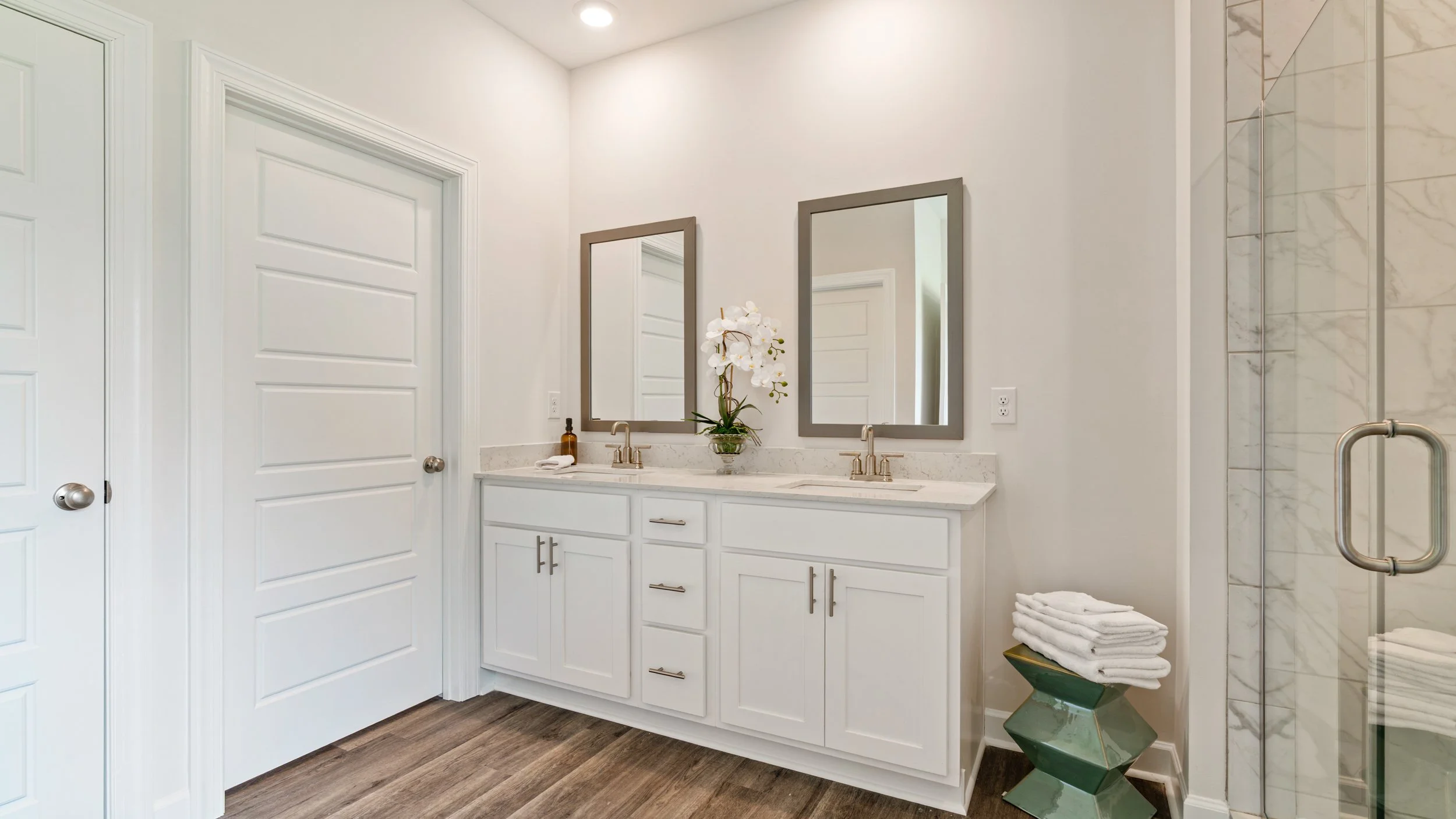 Modern bathroom with white double vanity, framed mirrors, white orchid plant, towels, wooden flooring, and a glass shower enclosure with marble tiles.