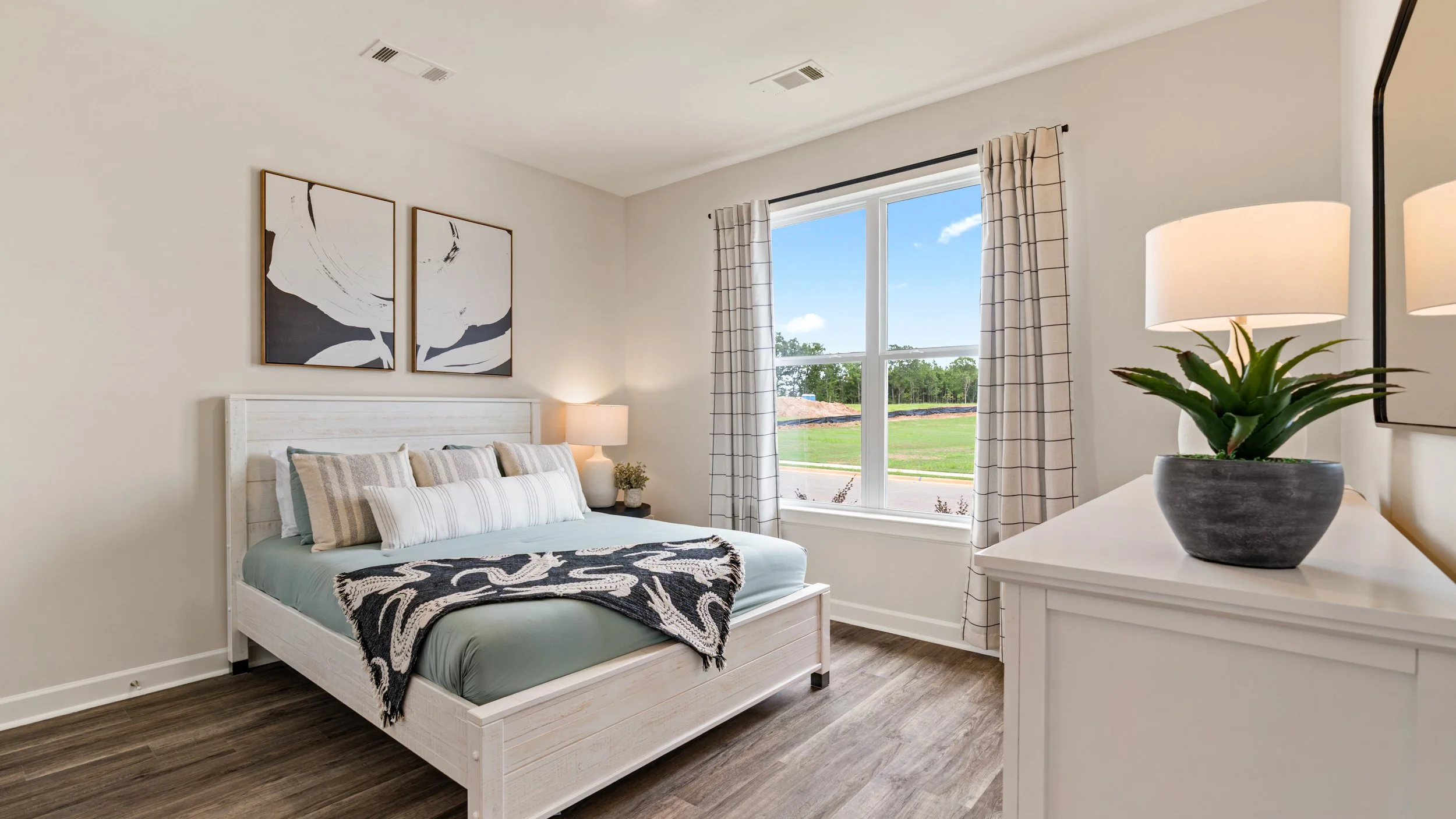 A bedroom with a white wooden bed, decorative pillows, a wall art above, a bedside lamp, a window with curtains, and a dresser with a potted plant.