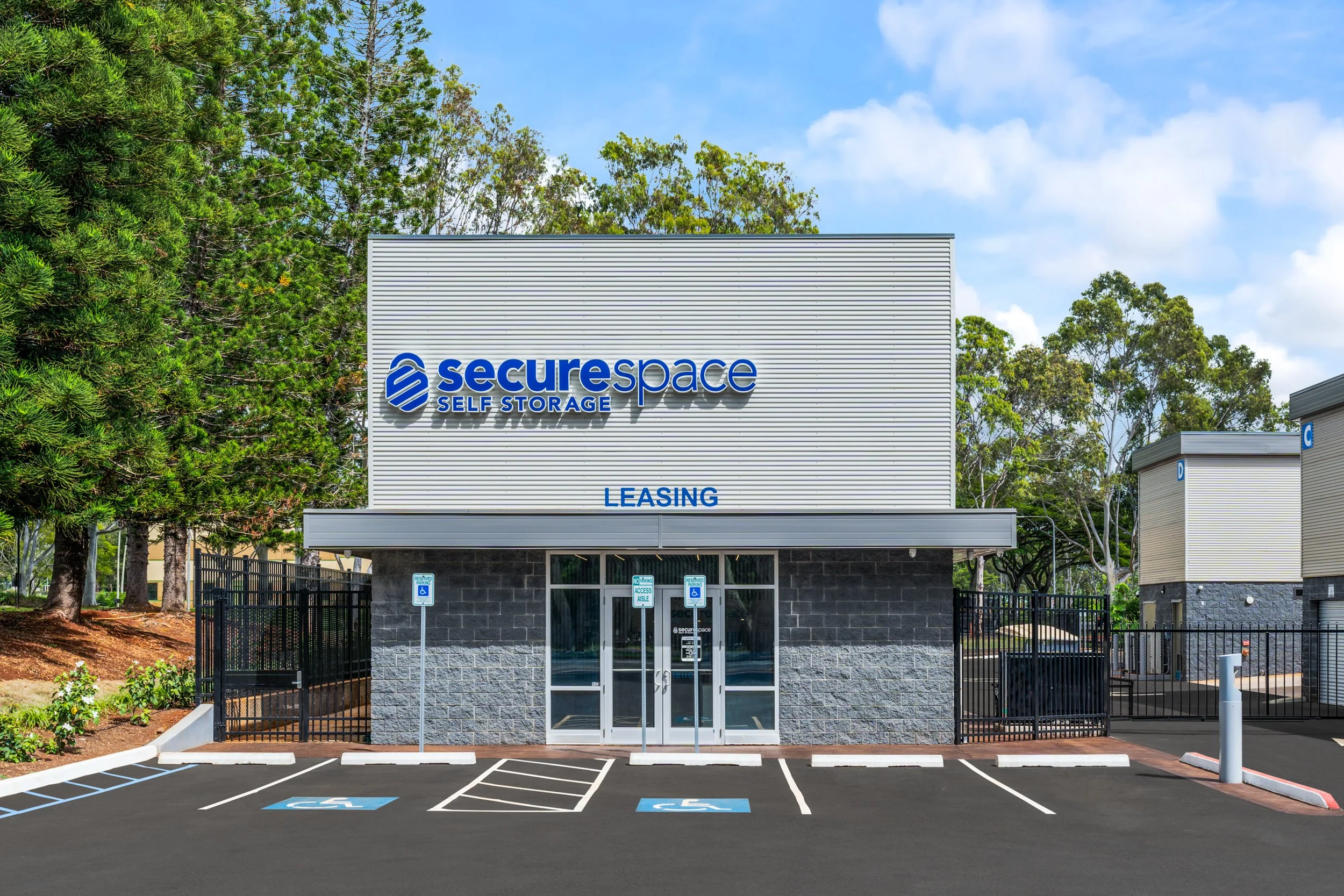 A self storage facility with the sign 'surespace Self Storage' and 'Leasing' written below. There are parking spaces in front, including designated handicapped spots, and an entrance with glass doors. Trees and a blue sky are visible in the backgroun