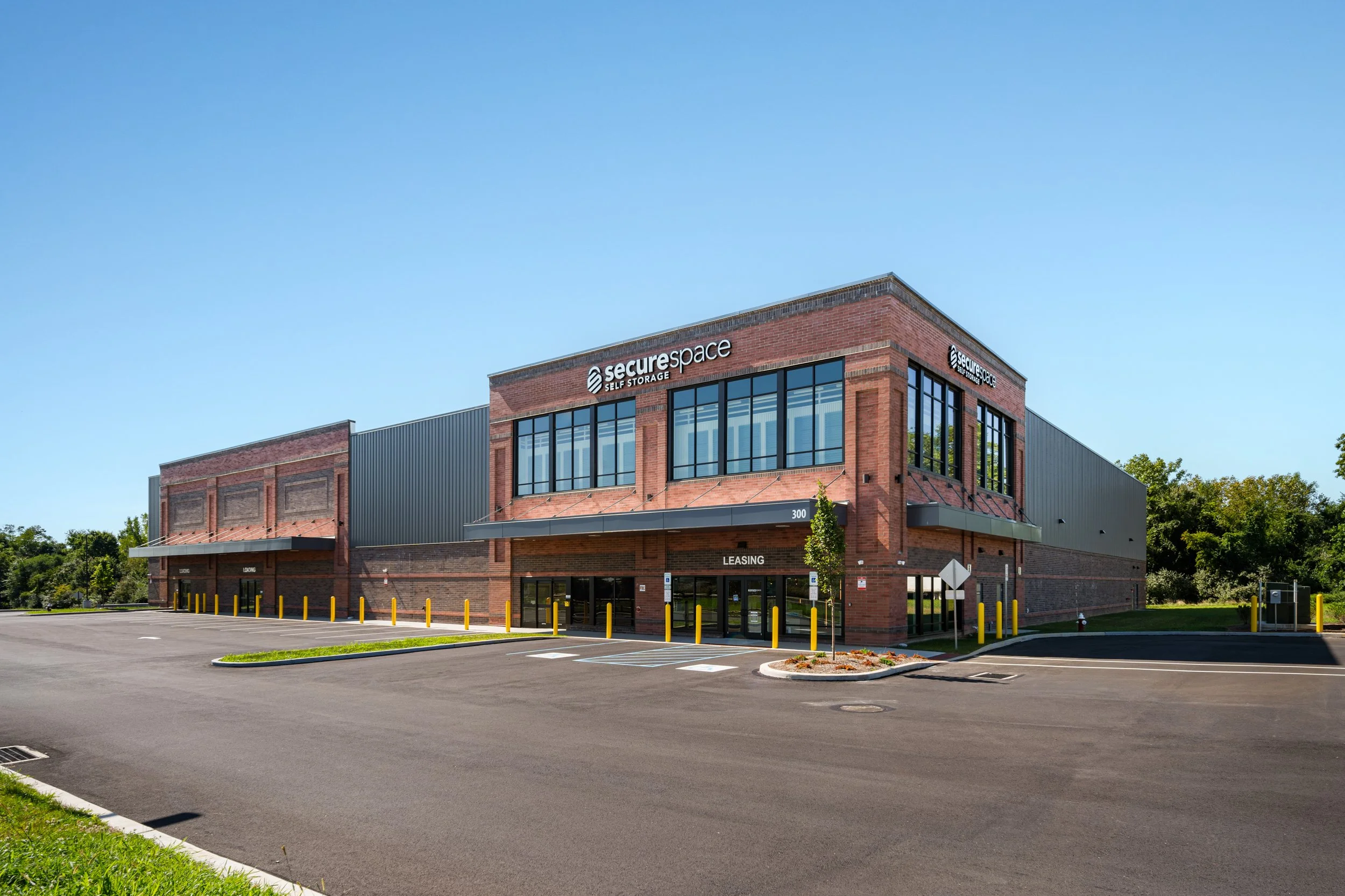Exterior view of a modern brick commercial building labeled 'SecureSpace Self Storage' with large glass windows, a parking lot, and a blue sky.