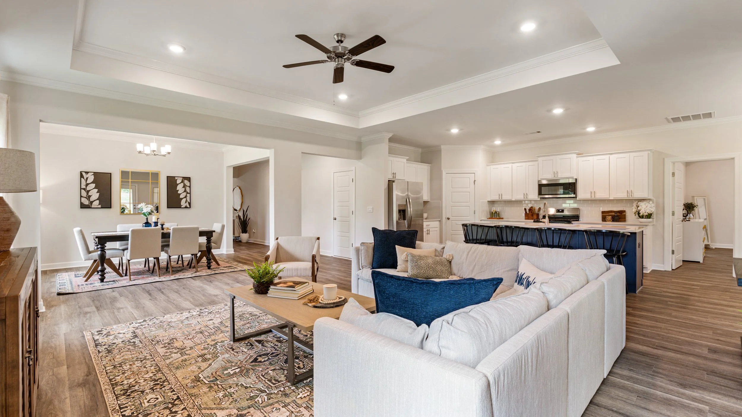 Open-concept living and dining area with white walls, hardwood floors, a white sectional sofa with beige and navy pillows, a wooden coffee table, a dining table with six white chairs, and a kitchen with white cabinets, a stainless steel refrigerator,