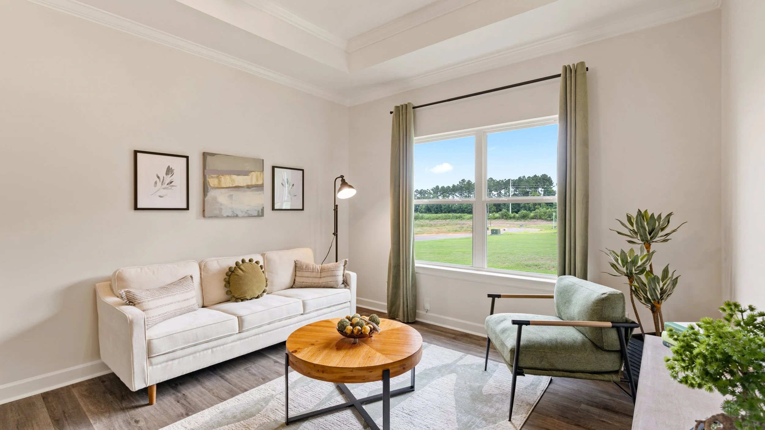 Living room with beige sofa, armchair, wooden coffee table, floor lamp, artwork on wall, window with green curtains, and plants.