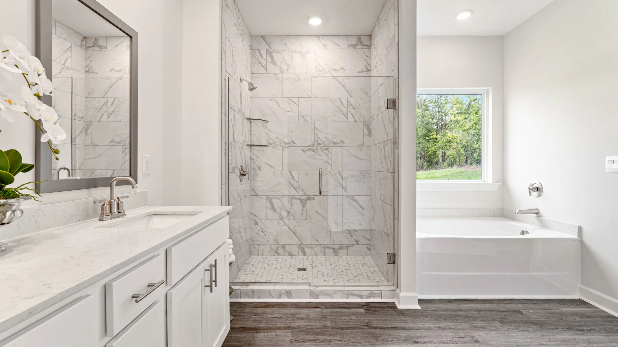 Bathroom with white vanity, marble countertop, large mirror, glass-enclosed walk-in shower with marble tiles, and a bathtub near a window showing greenery outside.