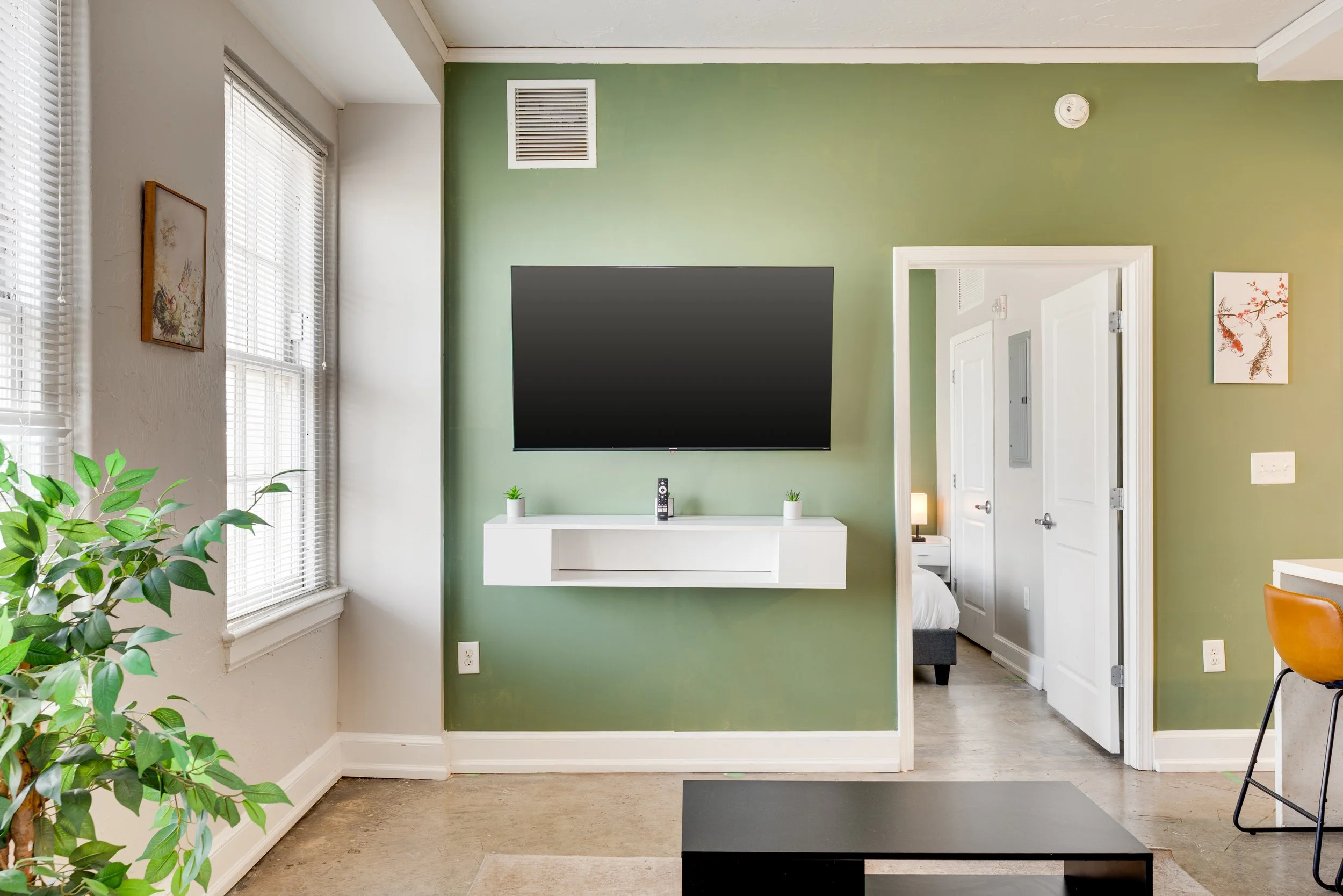 Living room with green accent wall, mounted flat-screen TV, white floating shelf, small potted plants, and a doorway leading to a bedroom.