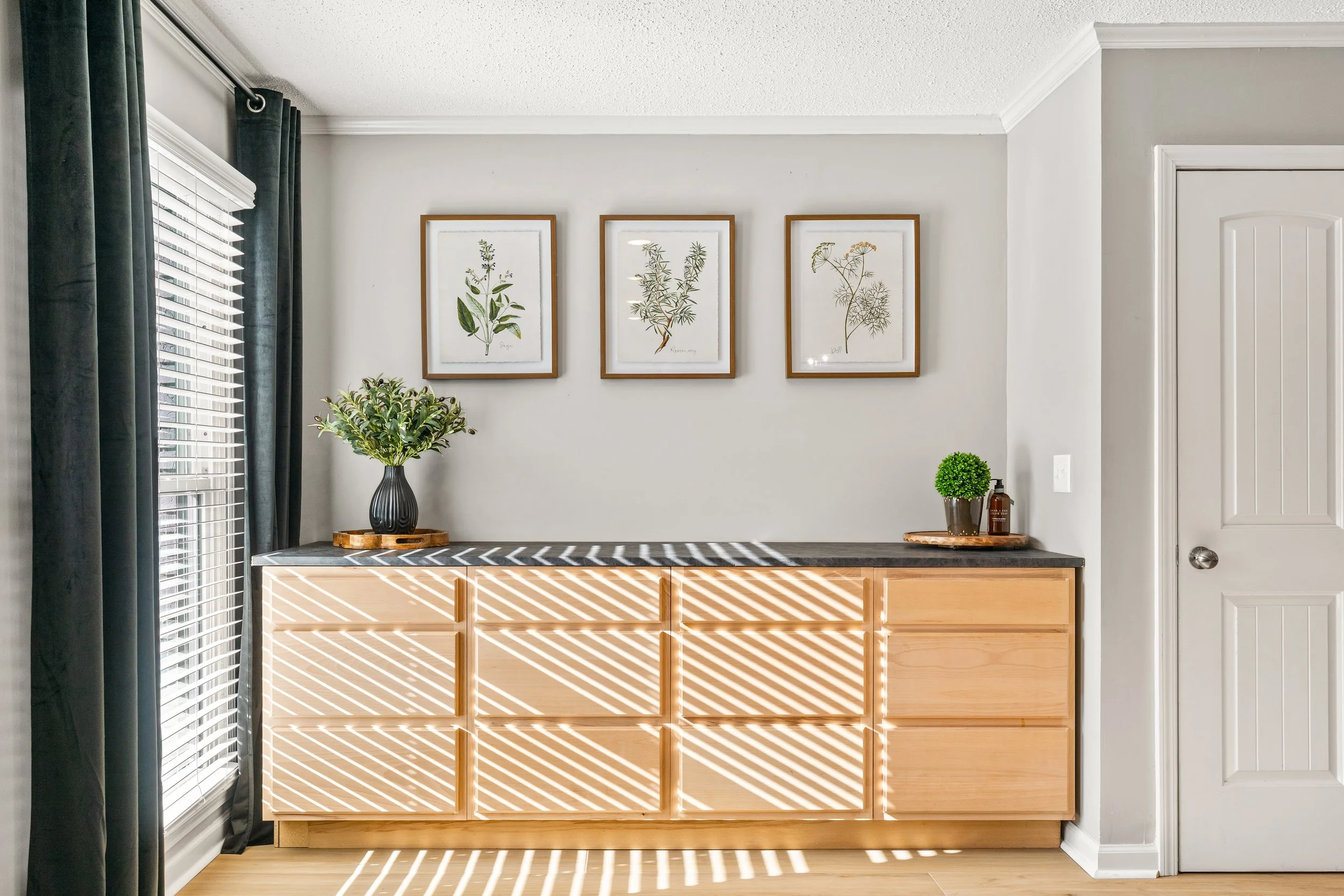 Interior of a room with a wooden sideboard, a black countertop, decorative plants, framed botanical prints, and sunlight filtering through blinds creating striped shadows.