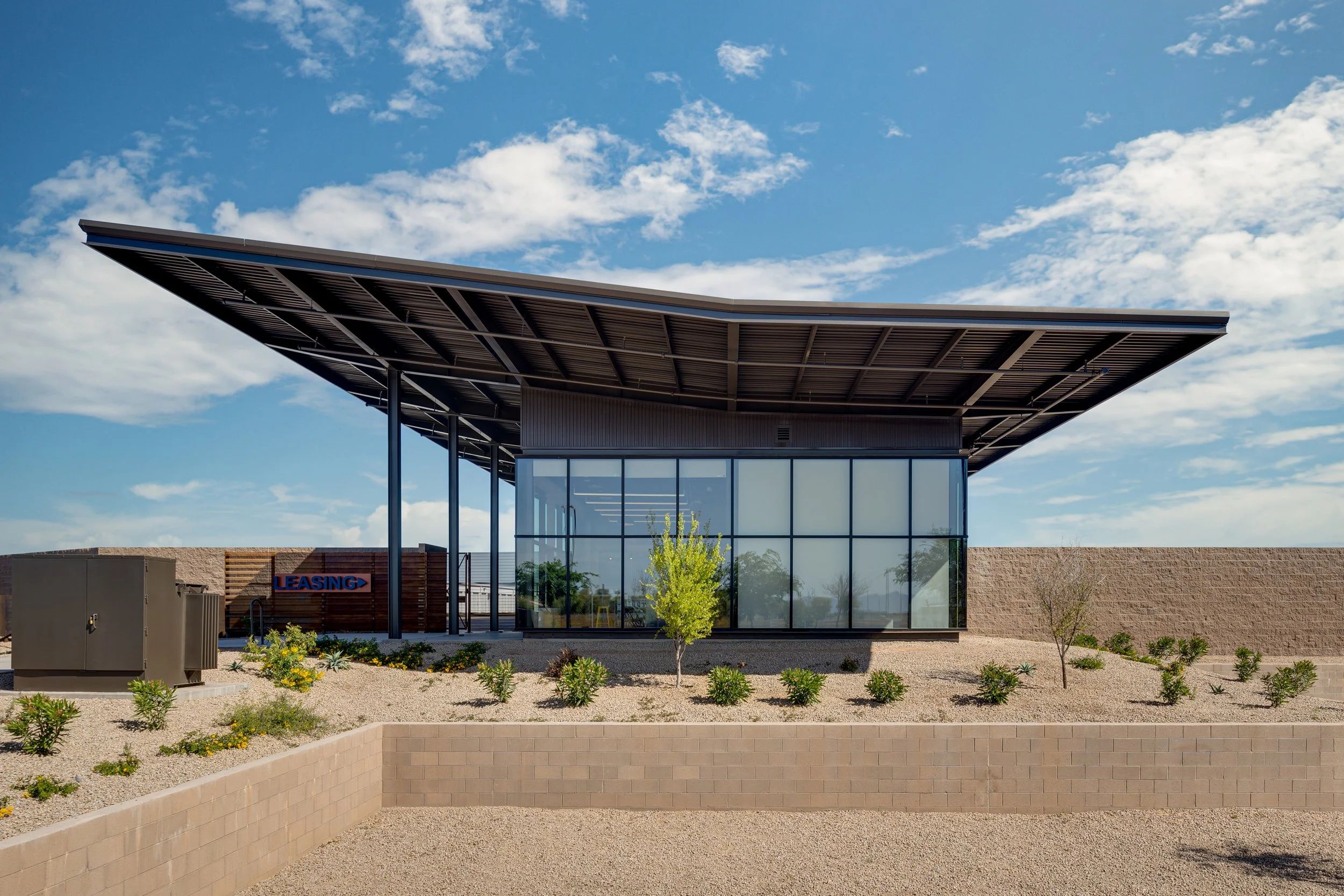 Modern office building with a distinctive angular roof and large glass windows, surrounded by desert landscaping with small bushes and trees, under a blue sky with clouds.