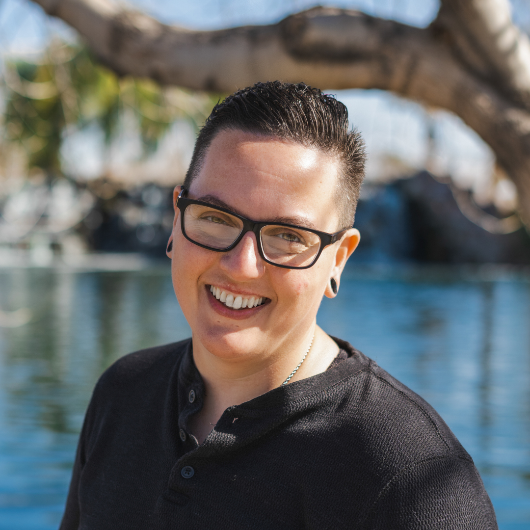 Smiling person with short dark hair, glasses, and gauges, outdoors near water with trees in the background.