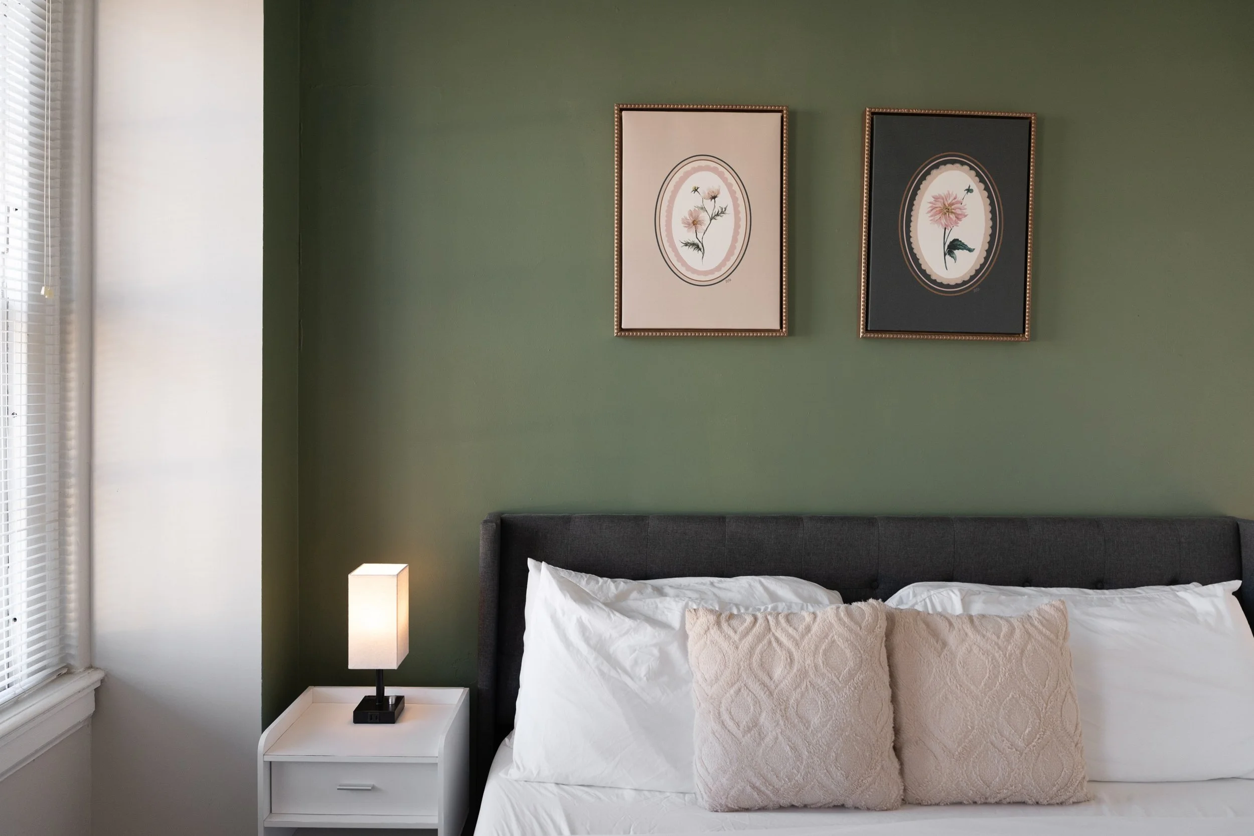 A bedroom with a dark gray upholstered headboard, white bedding, two decorative pillows, a white nightstand, a lamp, a window with blinds, and two framed botanical prints on a green wall.