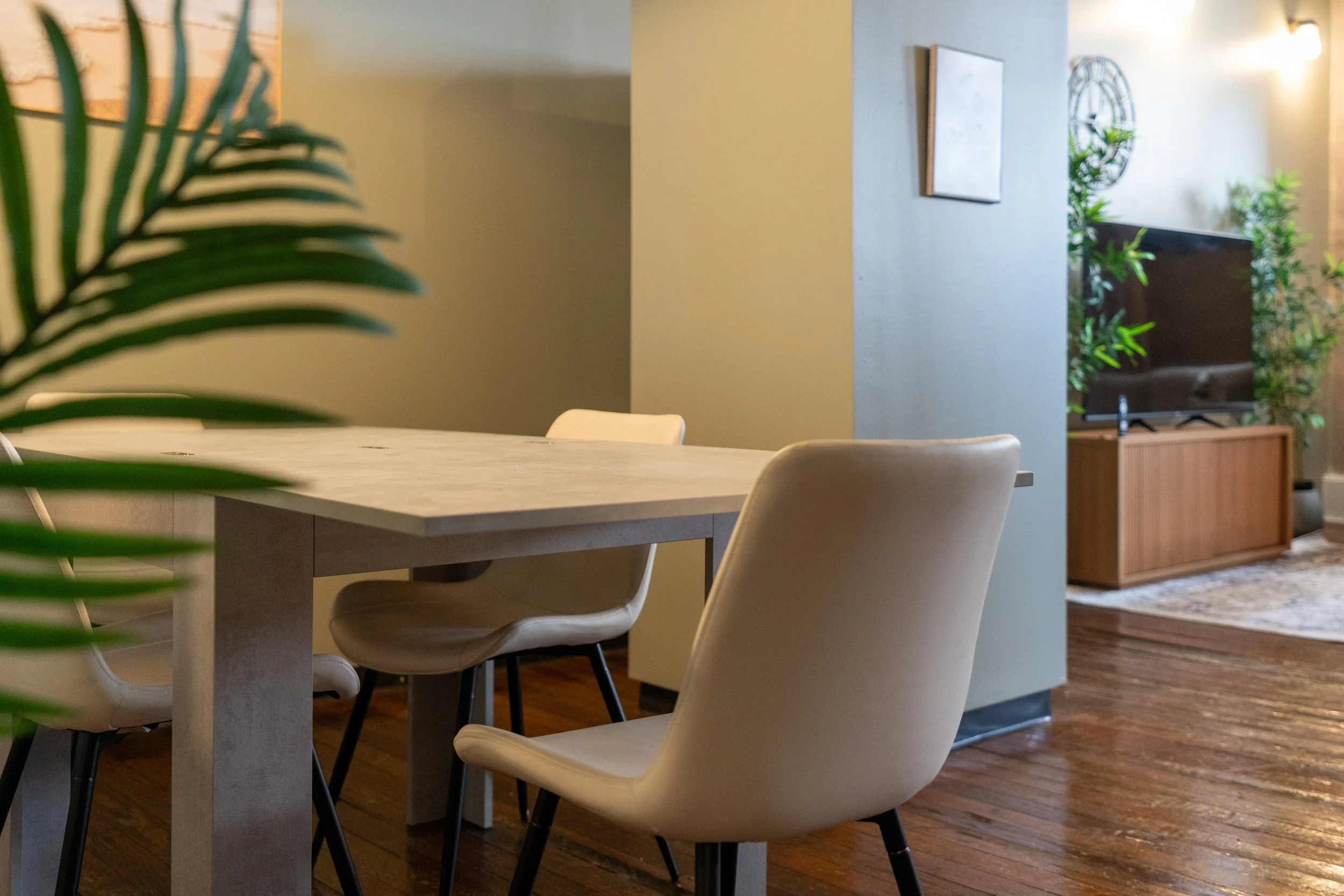 Dining area with a wooden table, beige chairs, and a living room with a TV and green plants in the background