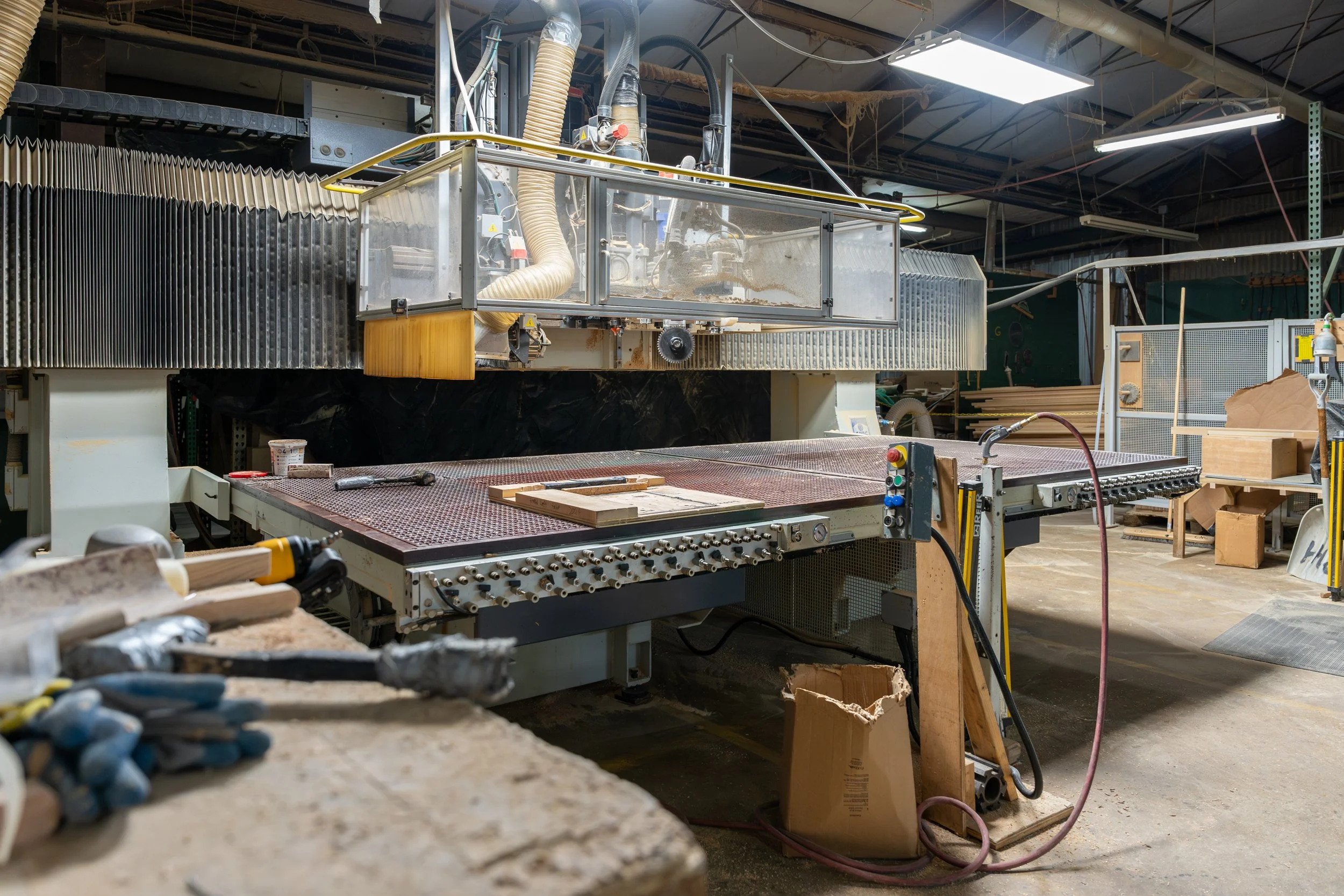 Industrial woodworking equipment in a workshop, including a large CNC machine, workbenches, tools, and assorted materials.