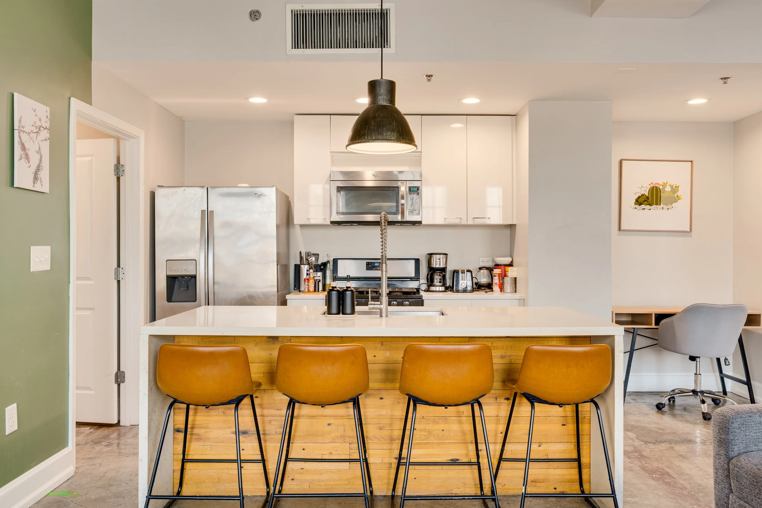 Modern kitchen with an island, four brown bar stools, stainless steel appliances, coffee maker, microwave, and small desk with gray chair.
