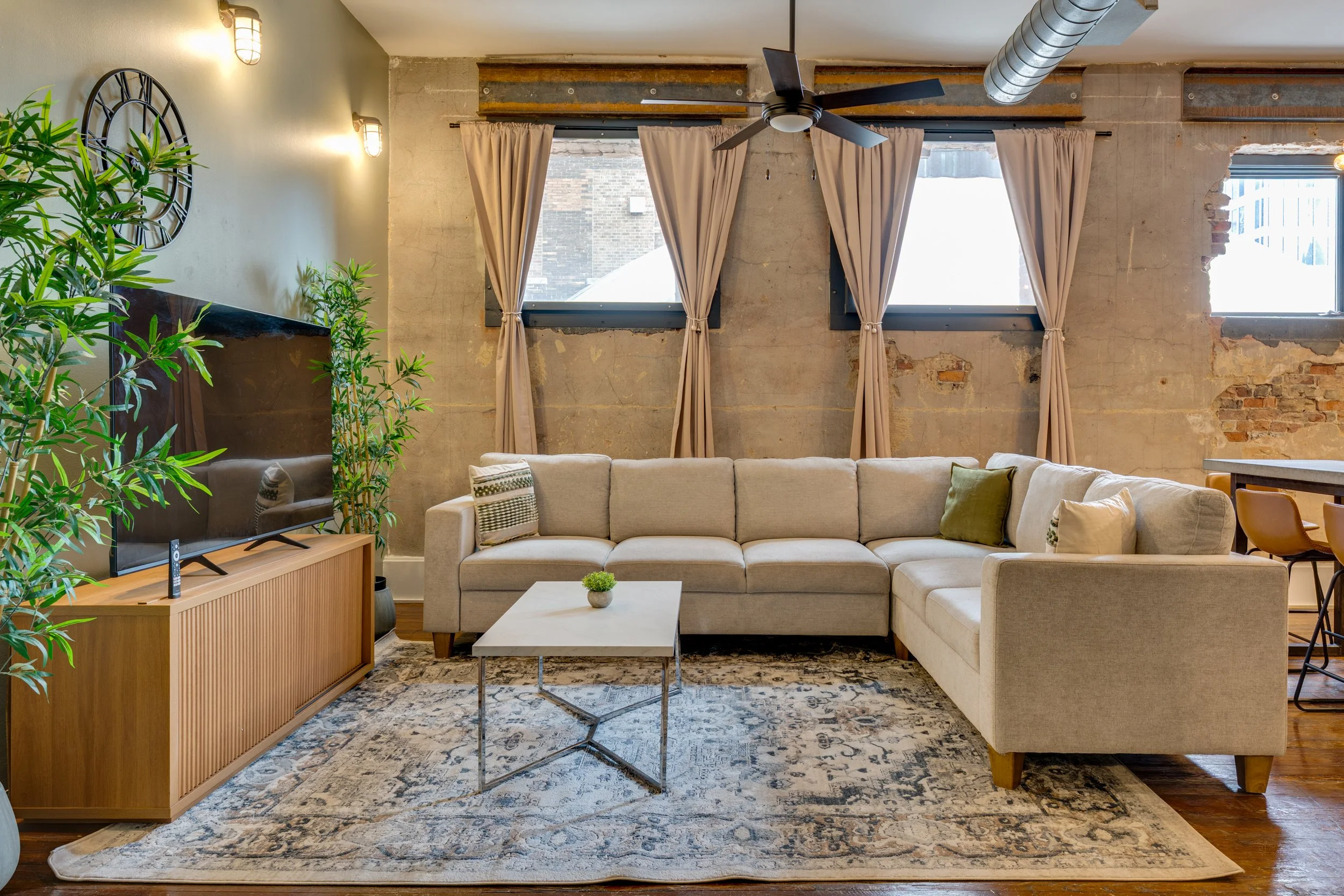 Modern living room with a beige sectional sofa, a white coffee table with a small plant, a large wall-mounted flat-screen TV, potted plants, and three windows with beige curtains, all in a space with exposed brick and industrial-style elements.
