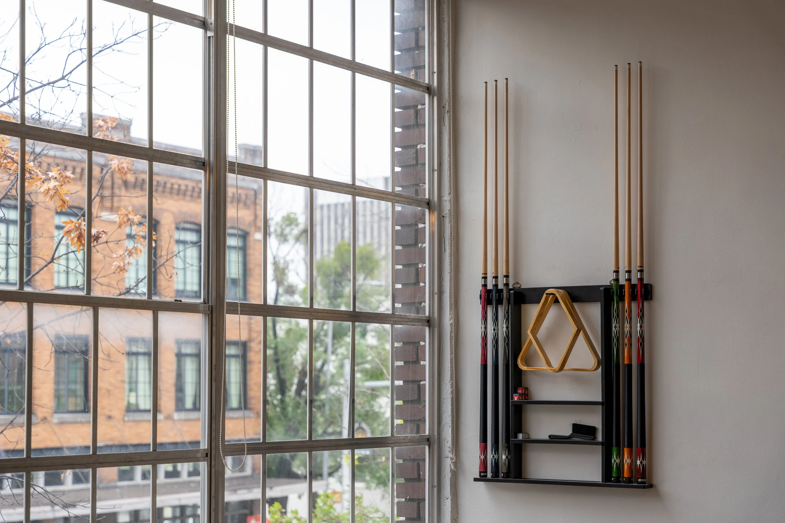 Indoor wall with pool cues stored vertically on a wall-mounted rack near a large window, with a view of buildings and trees outside.