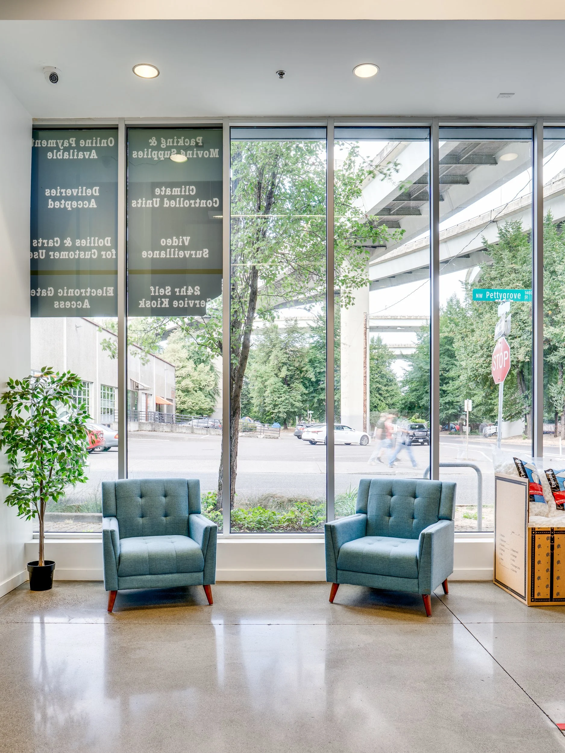 Interior view of a waiting area with two teal armchairs, a potted plant, and a large floor-to-ceiling window showing a street scene outside.