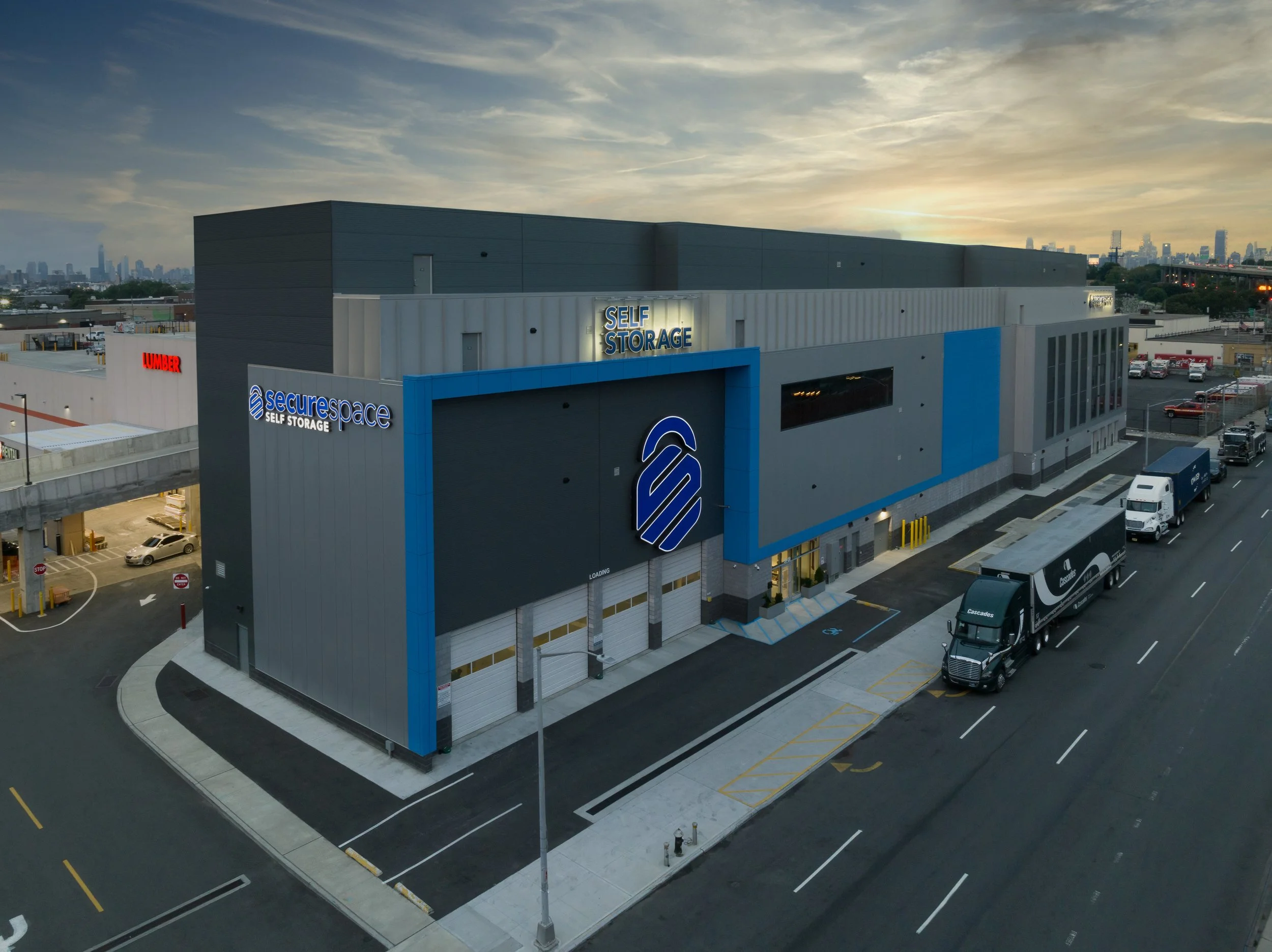 Exterior view of a secure self-storage facility with a modern gray and blue building, parking spaces, and trucks parked on the street, with a city skyline in the background during sunset.