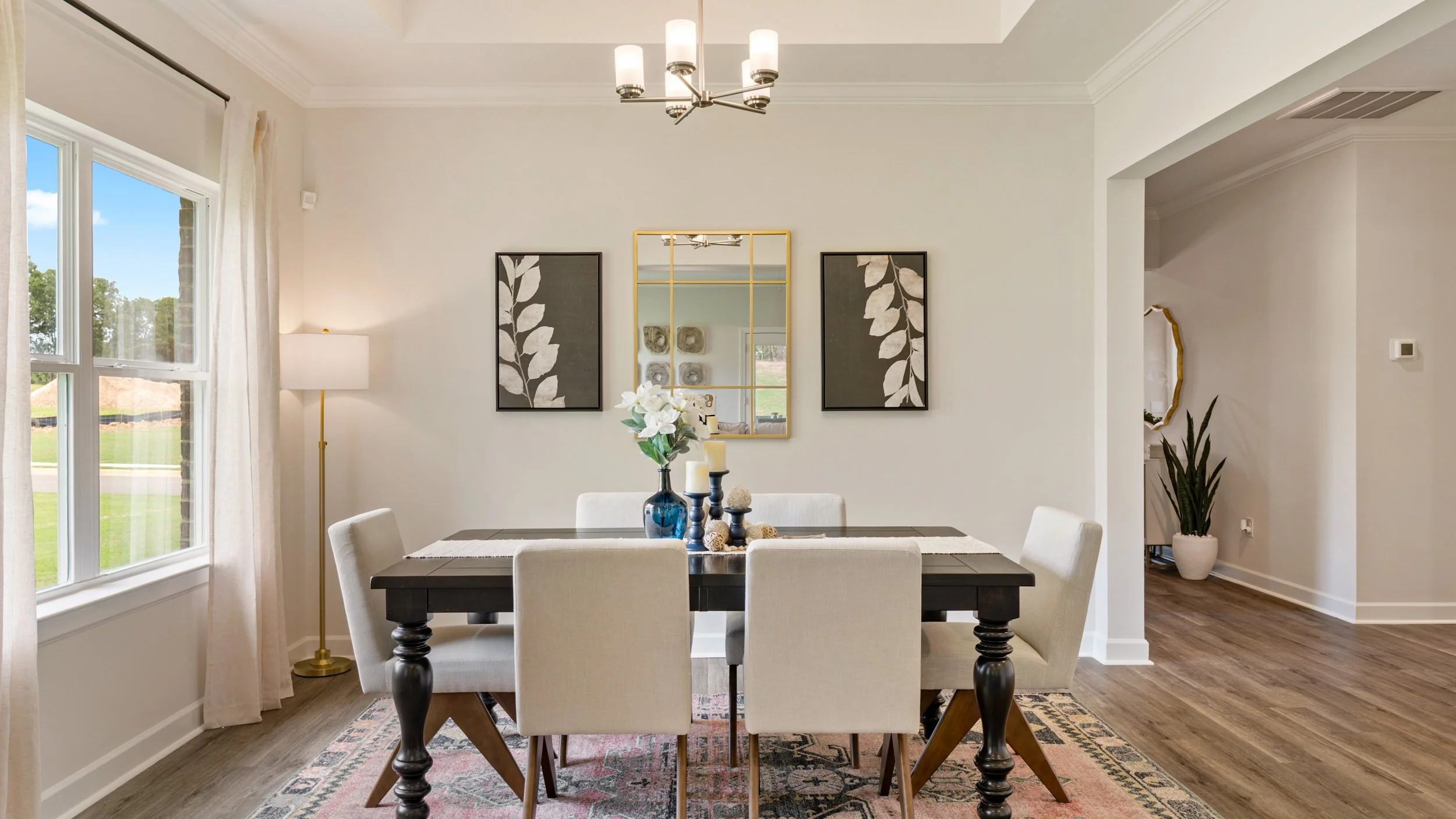 Dining room with a dark wooden table, six cream upholstered chairs, a blue vase with white flowers, decorative candles, a gold-framed mirror, two art prints of leaves, a large window with white curtains, a patterned rug, a floor lamp, and potted plan