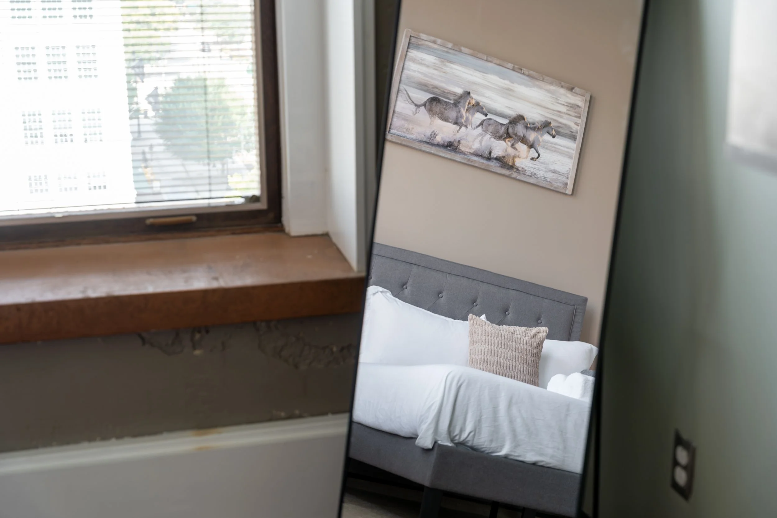 A split view showing a close-up of a window with blinds and a house wall on the left, and a bedroom with a gray upholstered bed and a large framed painting of horses on the right.