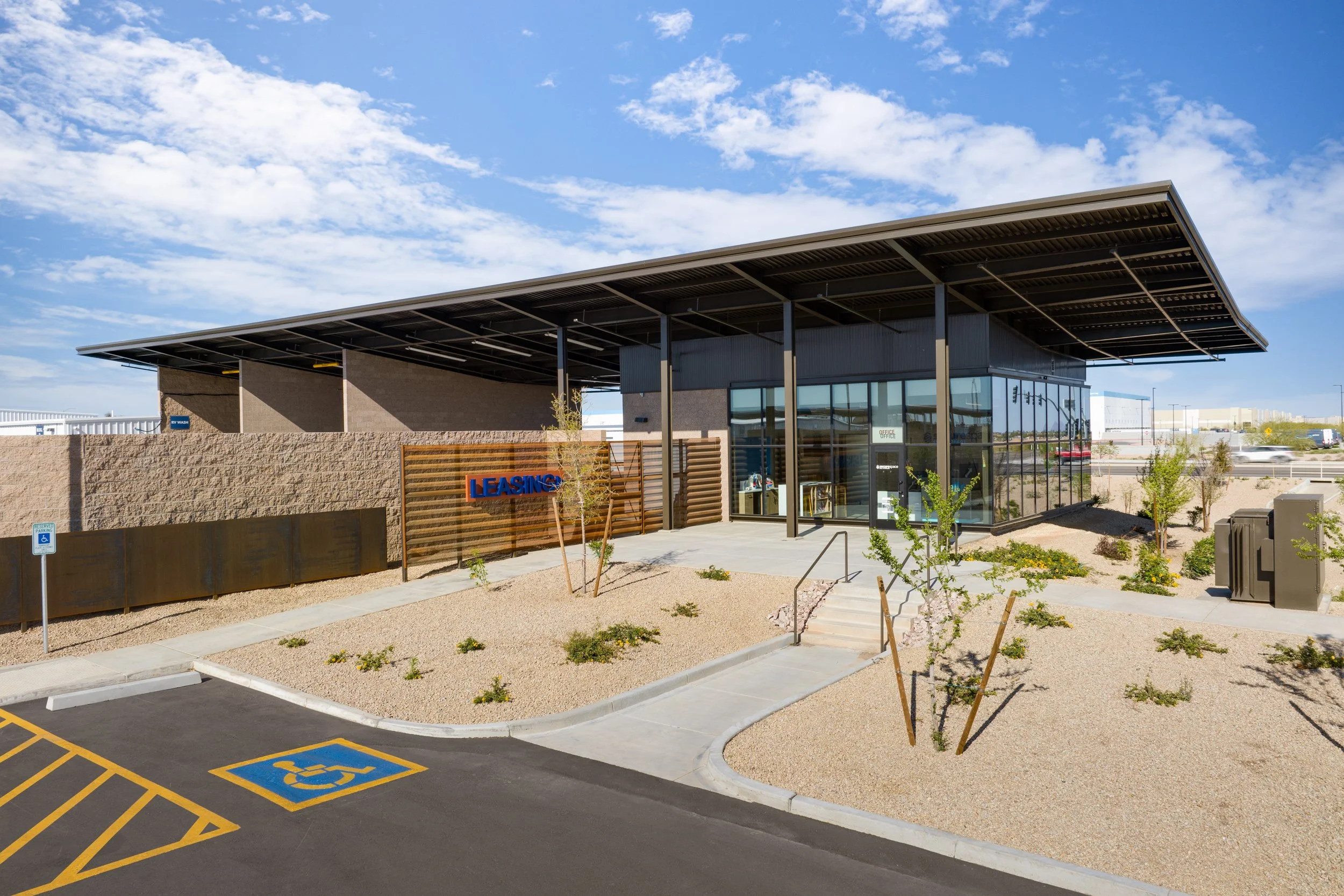 A modern fast food restaurant with a leasing sign, glass exterior, and a sloped metal roof, surrounded by a landscaped parking lot and small trees under a partly cloudy sky.