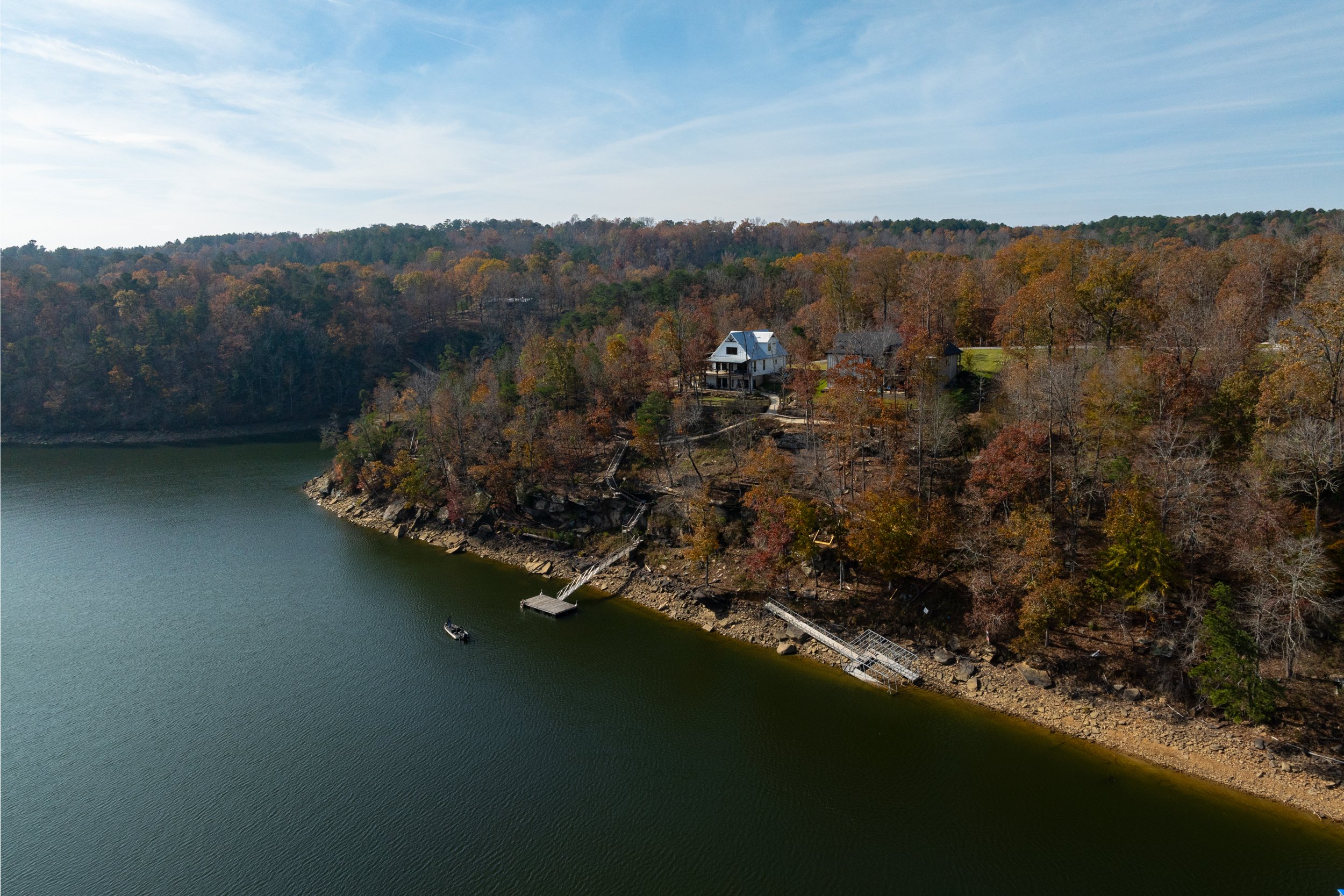 Aerial view of a lake with a dock, surrounded by wooded hills with houses, some trees showing fall colors.