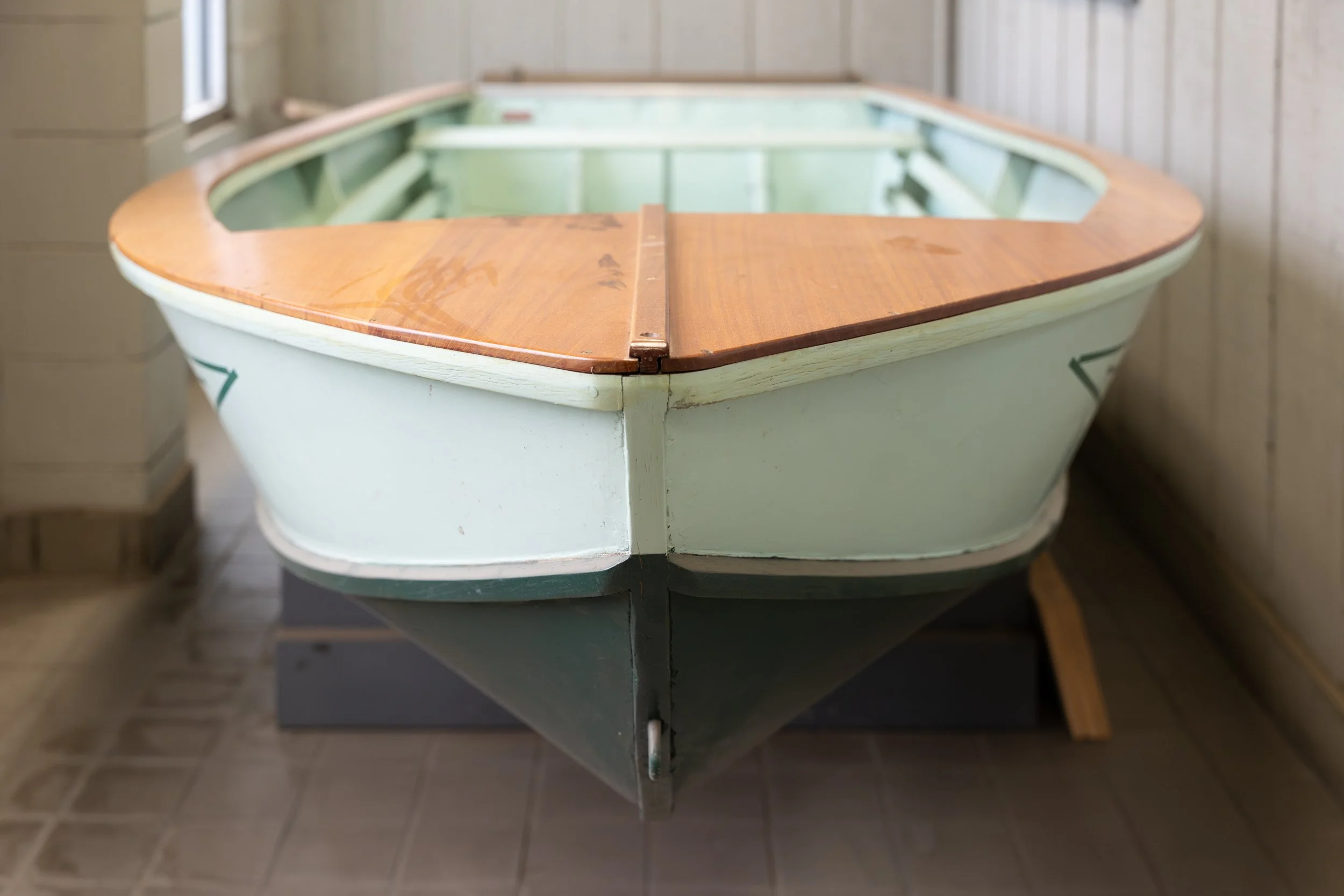 An old wooden boat with a light green hull and a brown wooden top, displayed indoors on a black stand with gray tiled floor and wood-paneled walls.
