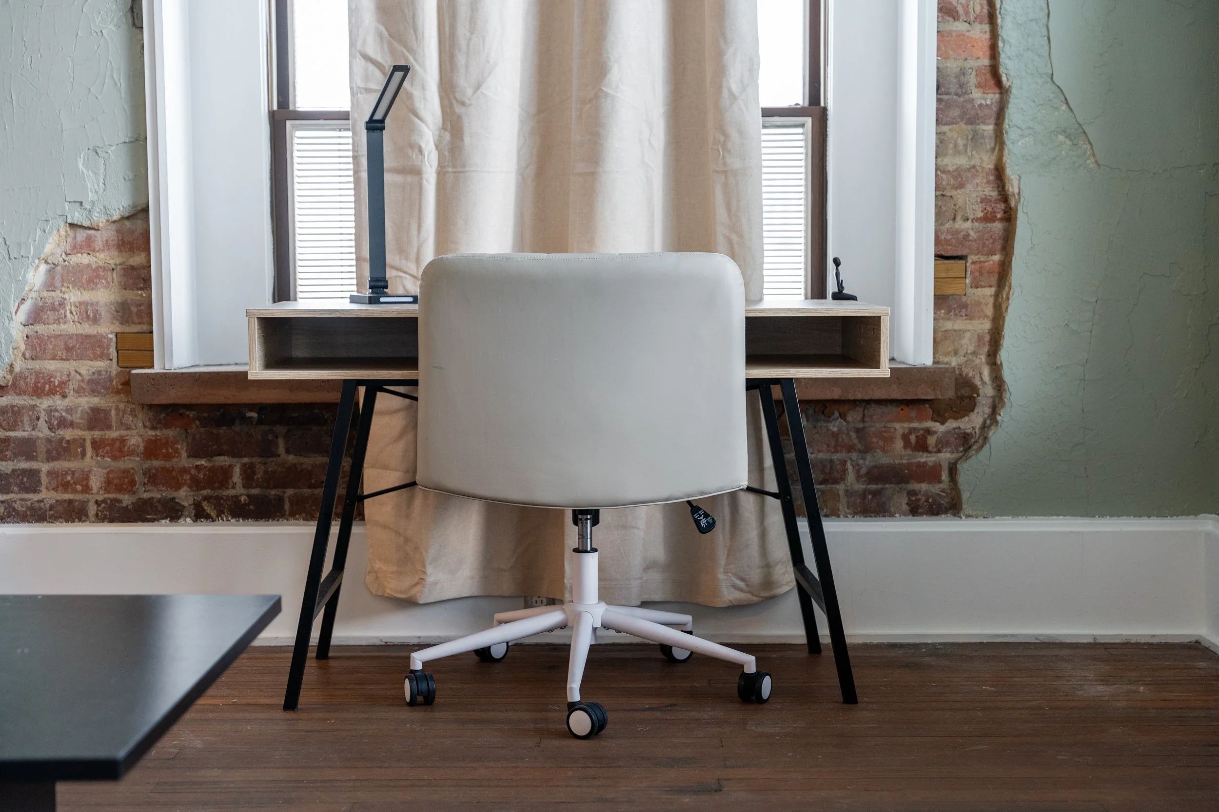 an office desk with a white chair in front of a window with beige curtains, exposed brick wall on one side, and a green wall on the other, hardwood floor