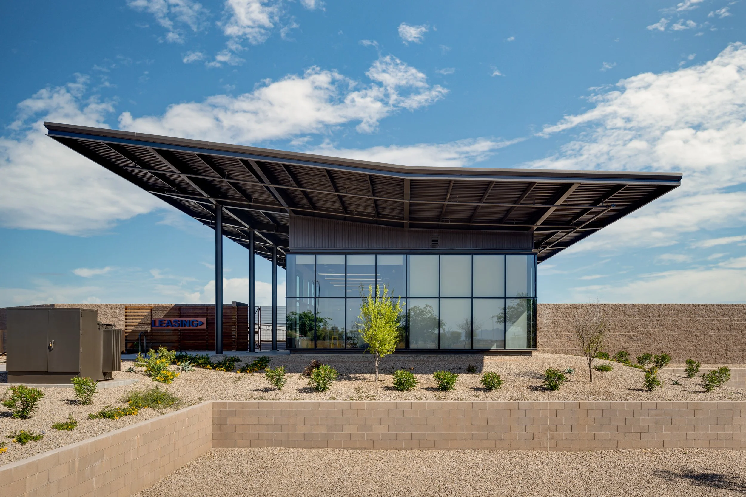 Modern commercial building with glass walls, a large sloped roof, and desert landscaping with small shrubs and trees.