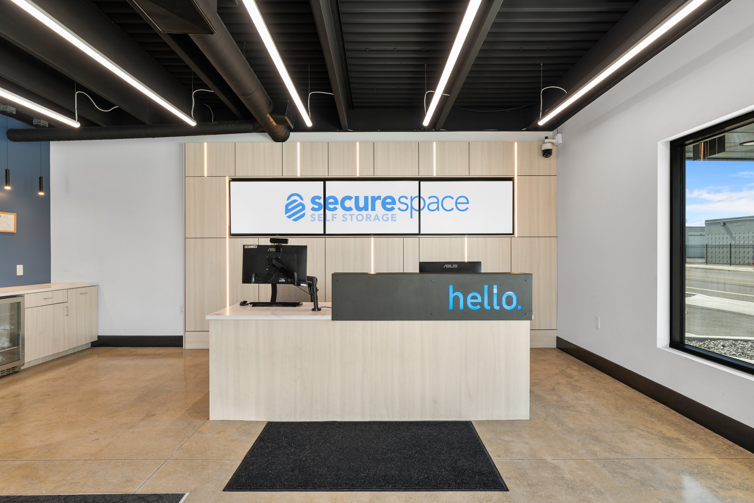 Reception area of a self-storage facility with a beige counter displaying the words 'hello.' in blue, a large screen behind with 'secure space SELF STORAGE' logo, two monitors on the counter, large window on right, and modern black ceiling with strip