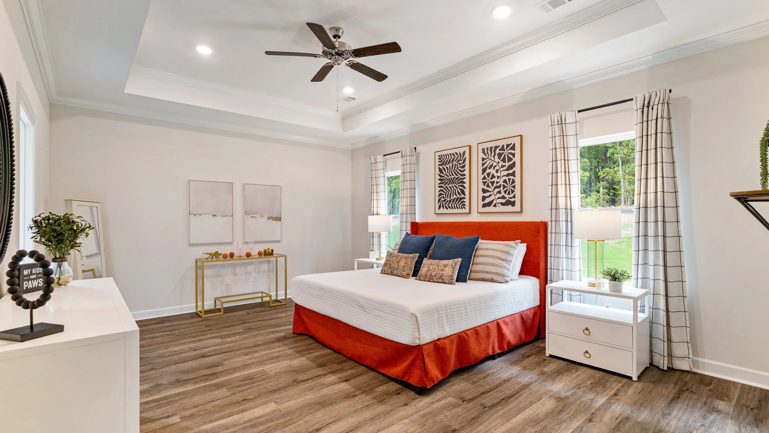 A modern bedroom with a white bed, orange headboard, dark blue and patterned pillows, white nightstands with lamps, large windows with checkered curtains, and interior decor including framed art and plants.