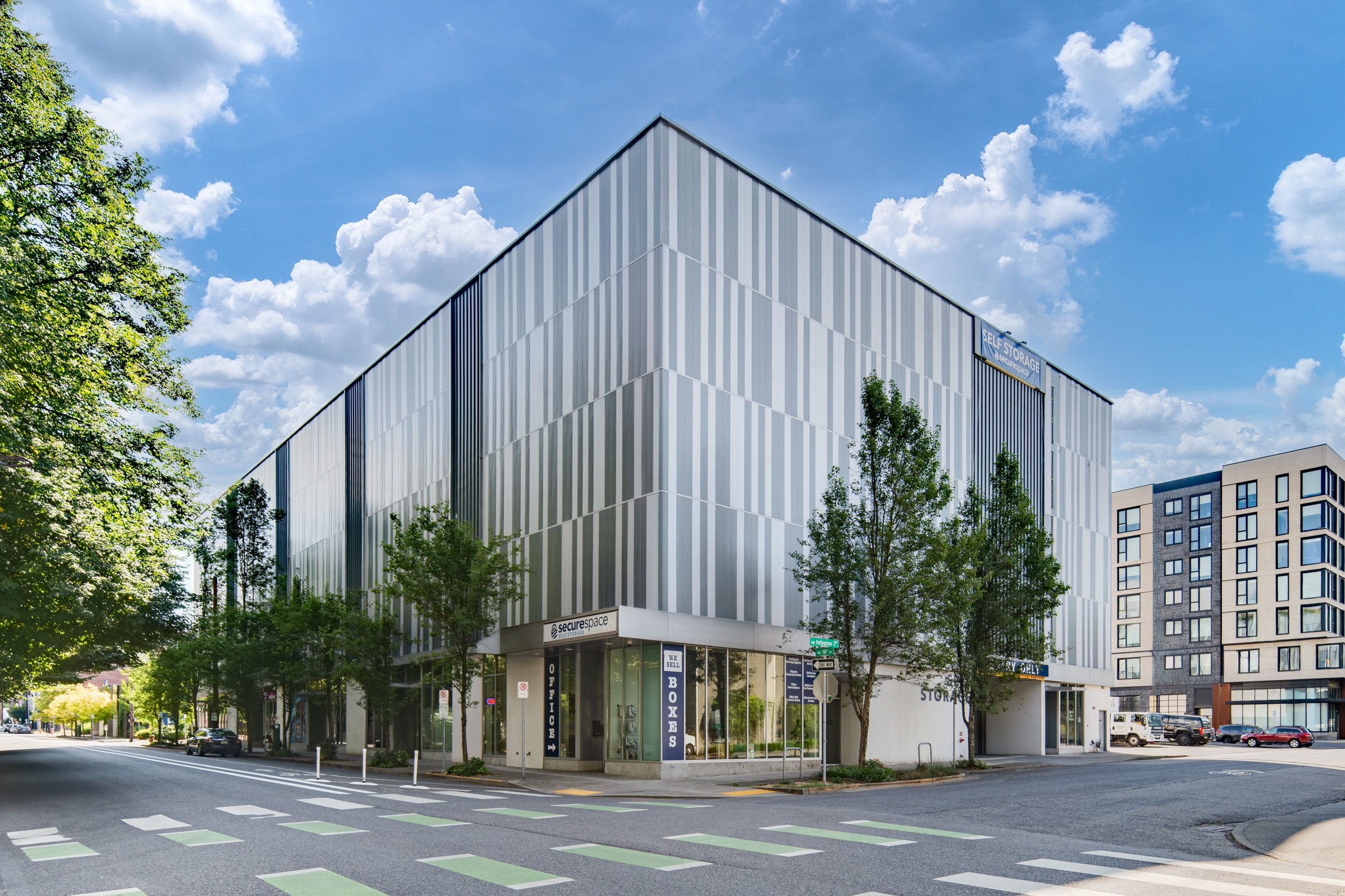 A modern multi-story commercial building with a sleek, metallic facade, situated on a city street corner under a partly cloudy sky, with trees and parked cars nearby.