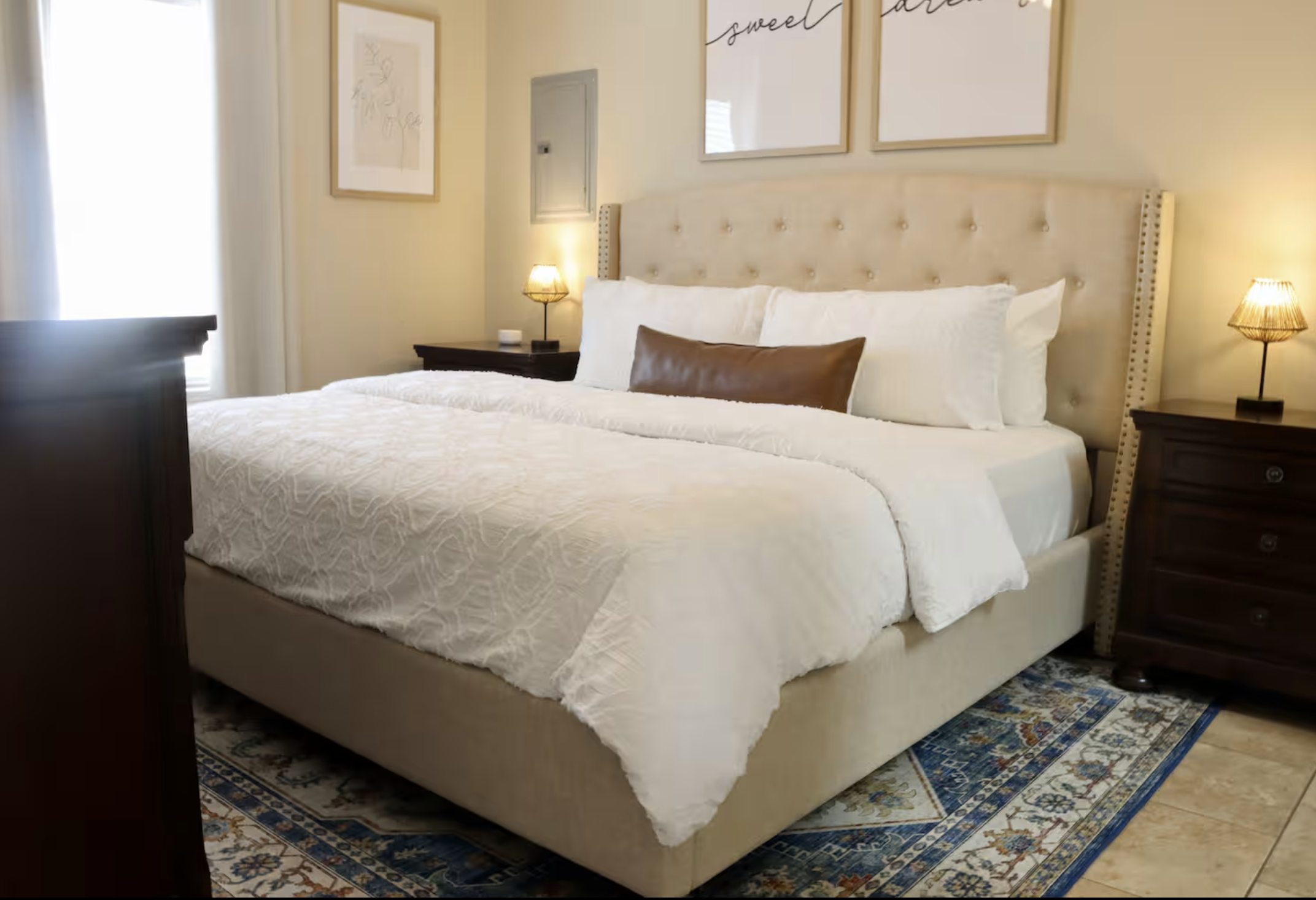 A neatly made bed with a beige upholstered headboard, white bedding, and a brown decorative pillow, located in a bedroom with framed wall art, bedside lamps, wooden nightstands, and a patterned rug.