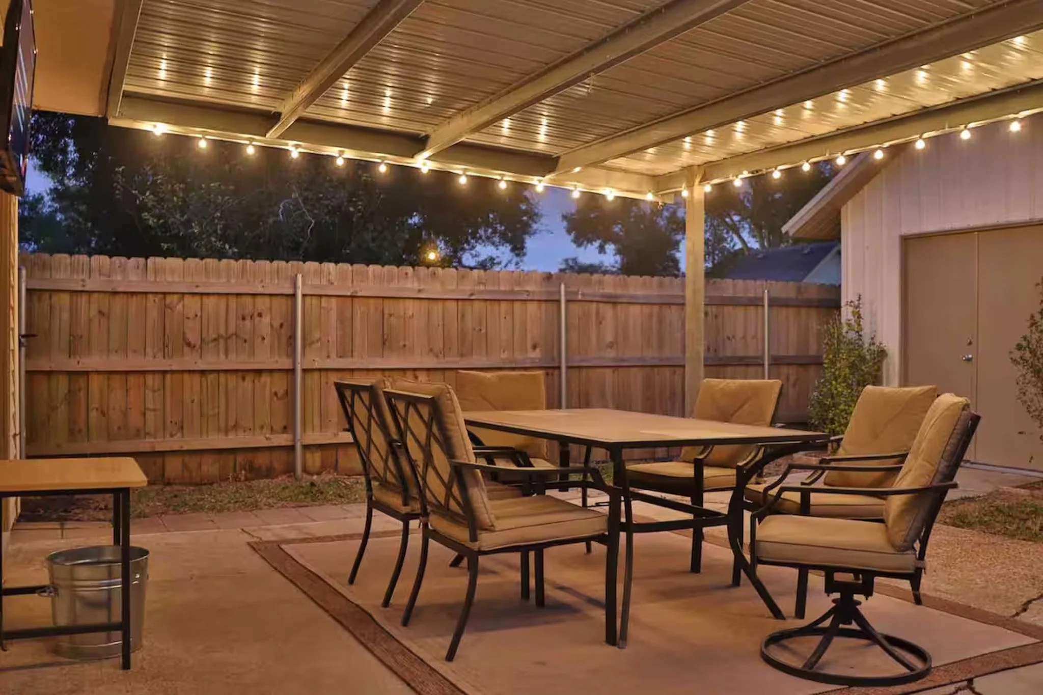 An outdoor patio area with a dining table and chairs, string lights hanging from a covered roof, a wooden fence in the background, and some plants beside the door.