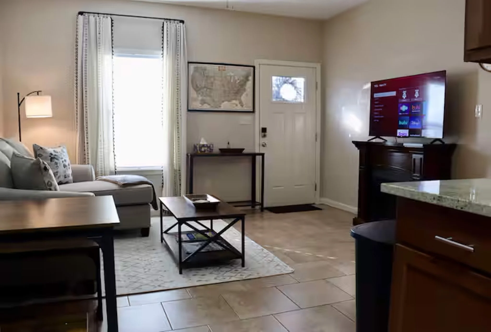 Living room with a beige sofa, patterned pillows, a wall lamp, a window with curtains, a small side table, a framed map on the wall, a door with a small window, a TV on a wooden stand, a marble countertop, and tile flooring.