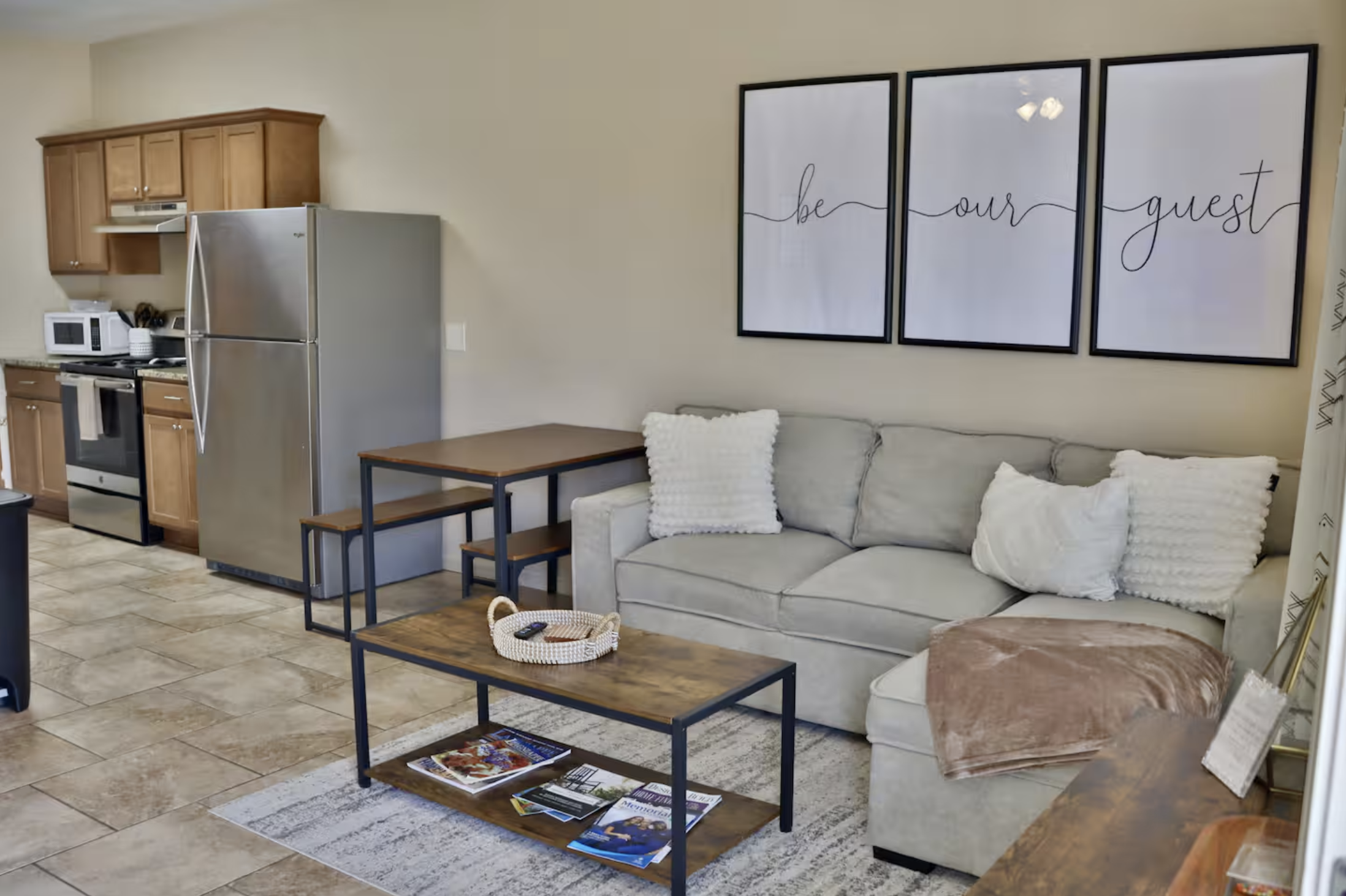 Living room area with a beige sofa decorated with white throw pillows, a coffee table with magazines and a remote, and wall art that reads 'be our guest'. The open kitchen has wooden cabinets, a stainless steel refrigerator, microwave, stove, and small dining table with benches.