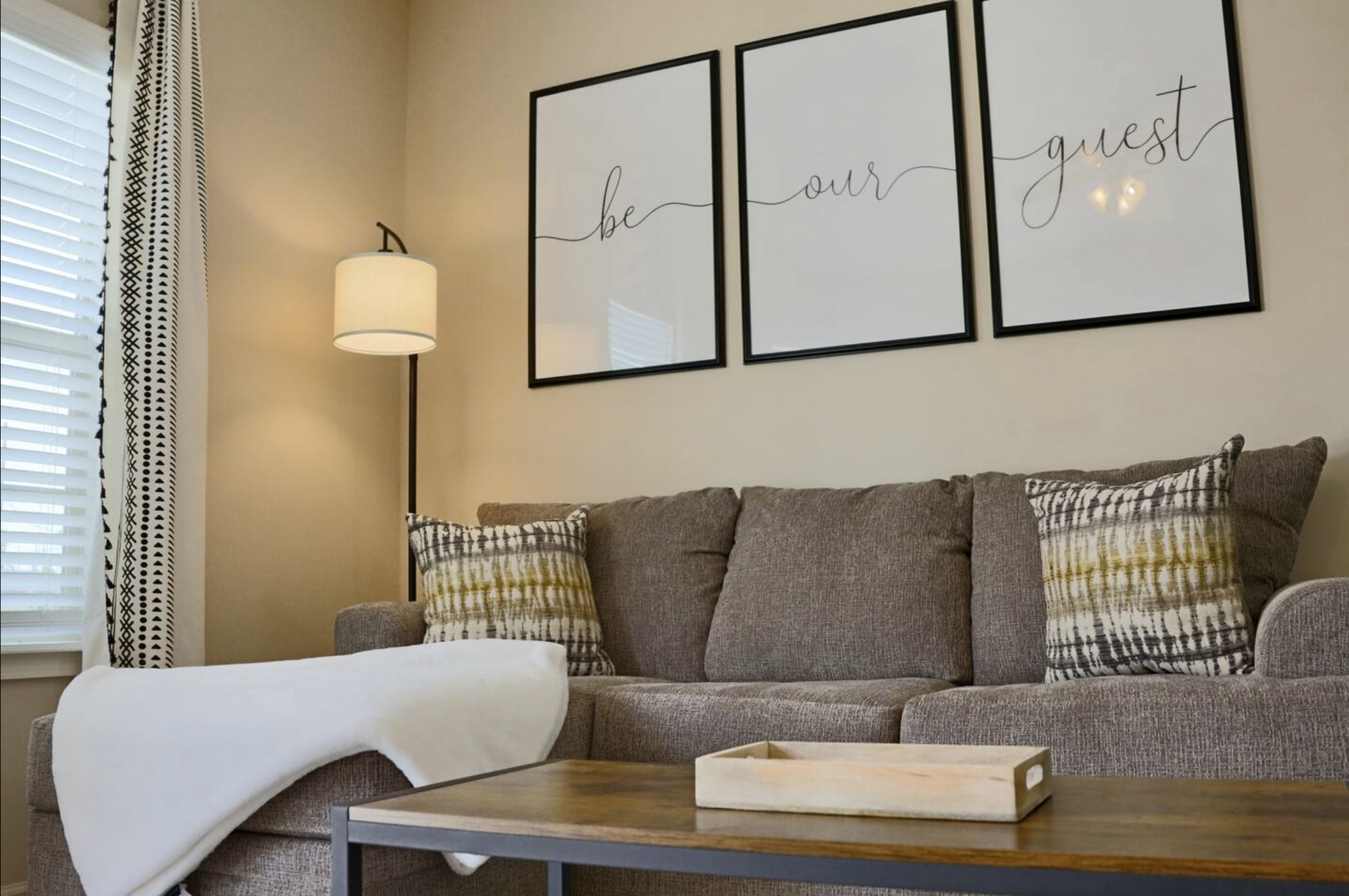 Living room with beige wall, framed art saying 'be our guest', gray sofa with patterned pillows, white throw blanket, wooden coffee table with tray, window with black and white curtains, floor lamp with white shade.