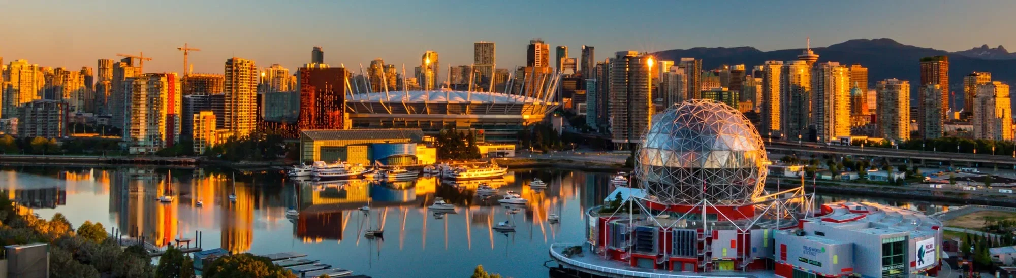 City skyline view of Vancouver with tall buildings, a stadium, and the Science World geodesic dome reflecting in the water at sunset.