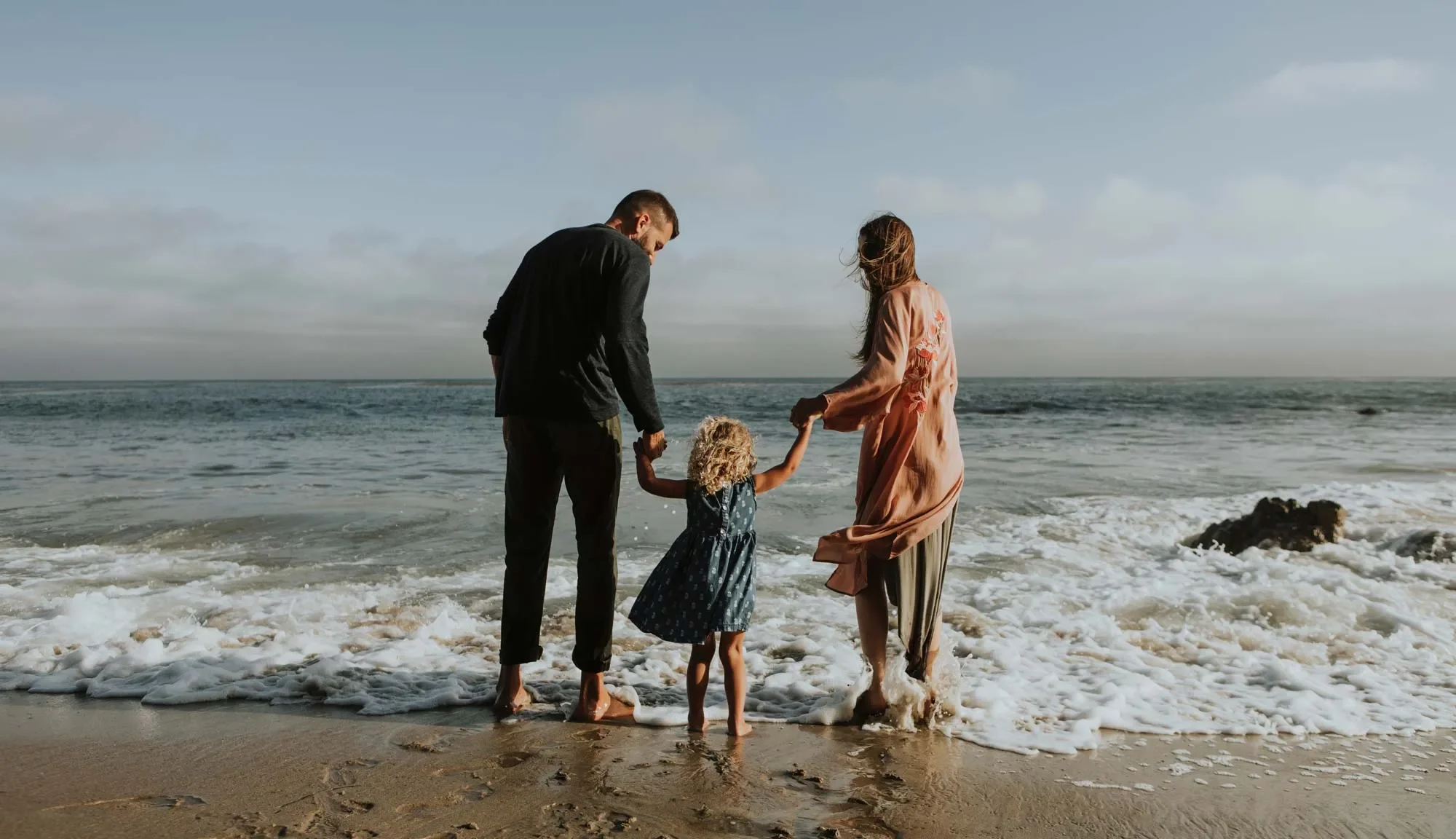 A family of three standing at the shoreline, holding hands, with a man and woman on either side of a young girl, all facing the ocean as waves lap at their feet on a sandy beach under a partly cloudy sky.