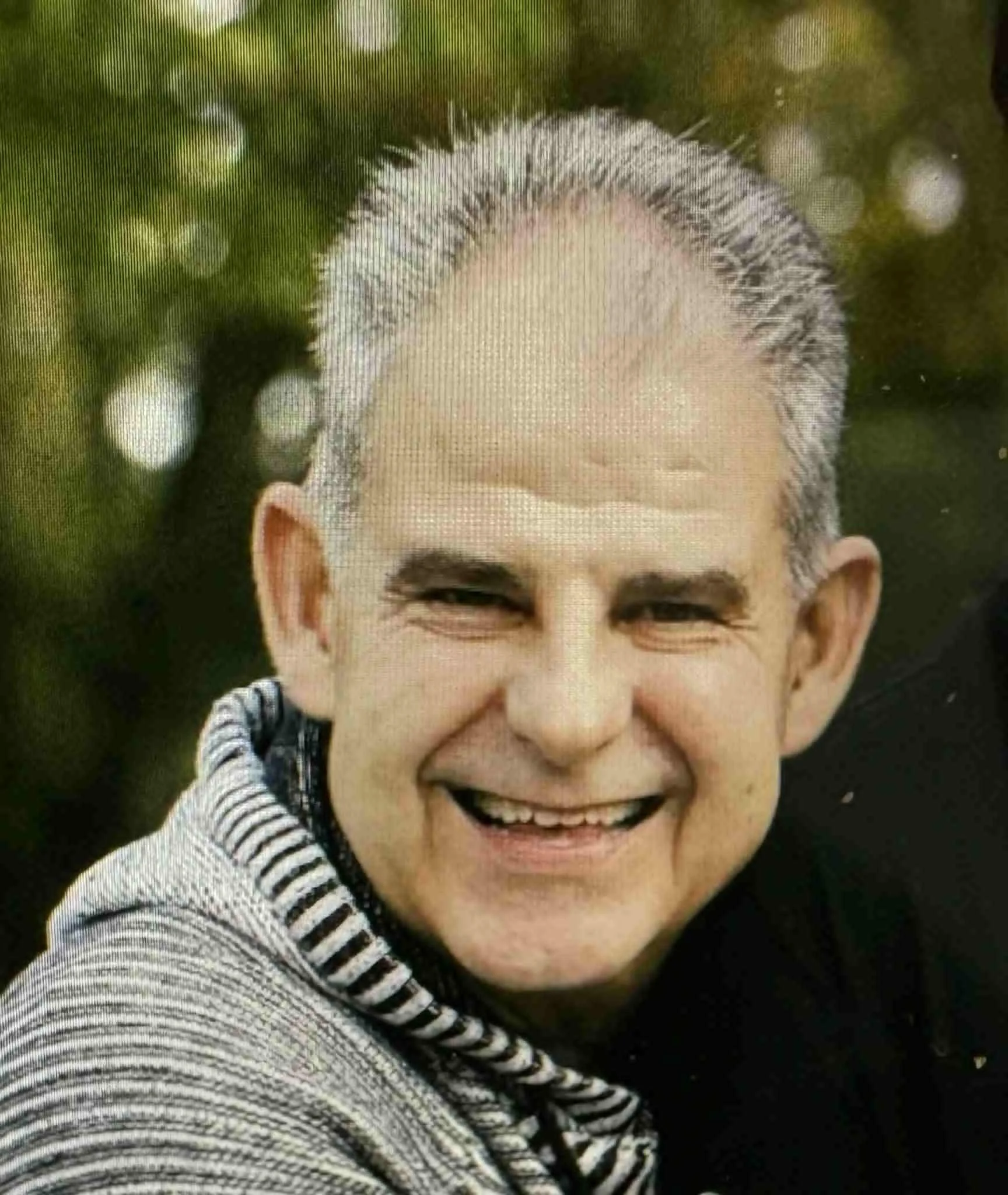 Close-up of a smiling man with short gray hair, wearing a black shirt and striped sweater, outdoors with green bokeh background.