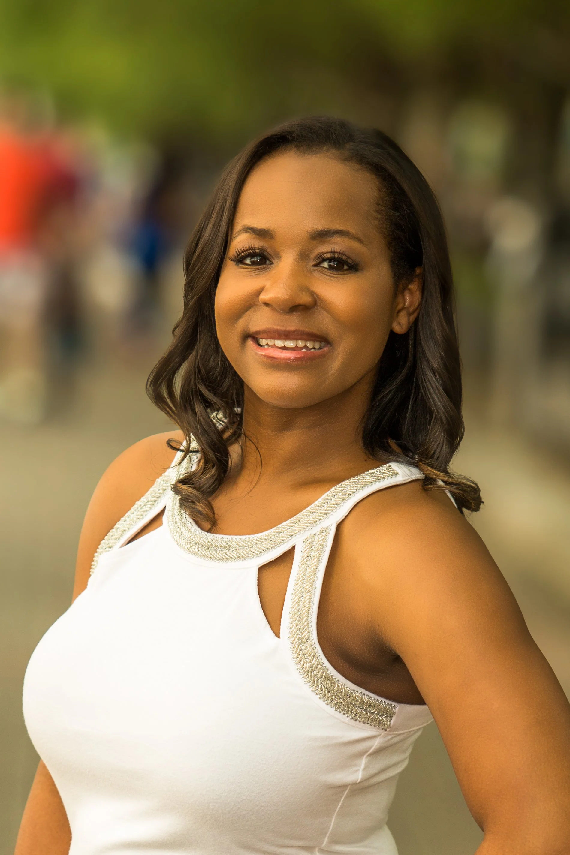 Portrait of a woman with shoulder-length dark hair, smiling, wearing a white sleeveless top with embellished neckline, standing outdoors with a blurred background.