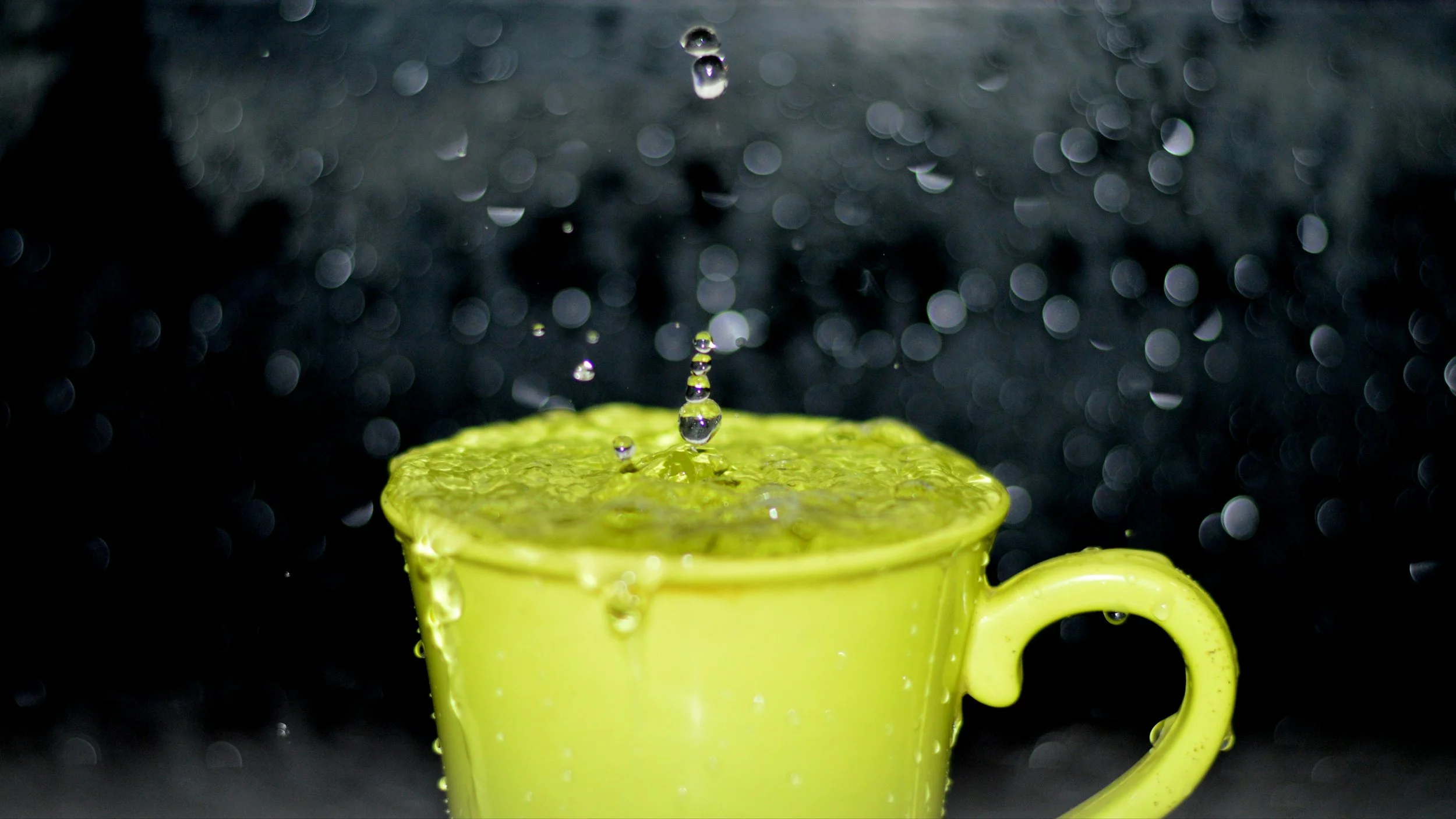 A yellow cup overflowing with water as a single drop lands on the surface, representing how one small addition to an already full sprint can break a product team's capacity and focus.